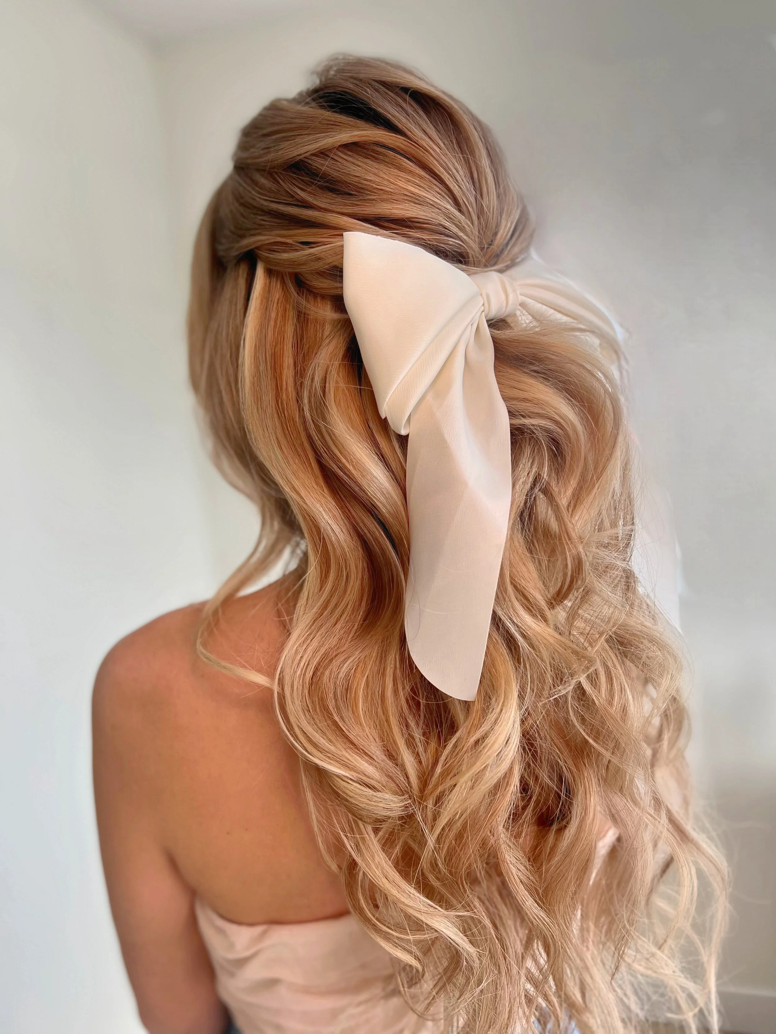 A woman with long, wavy blonde hair tied with a large cream-colored satin bow.
