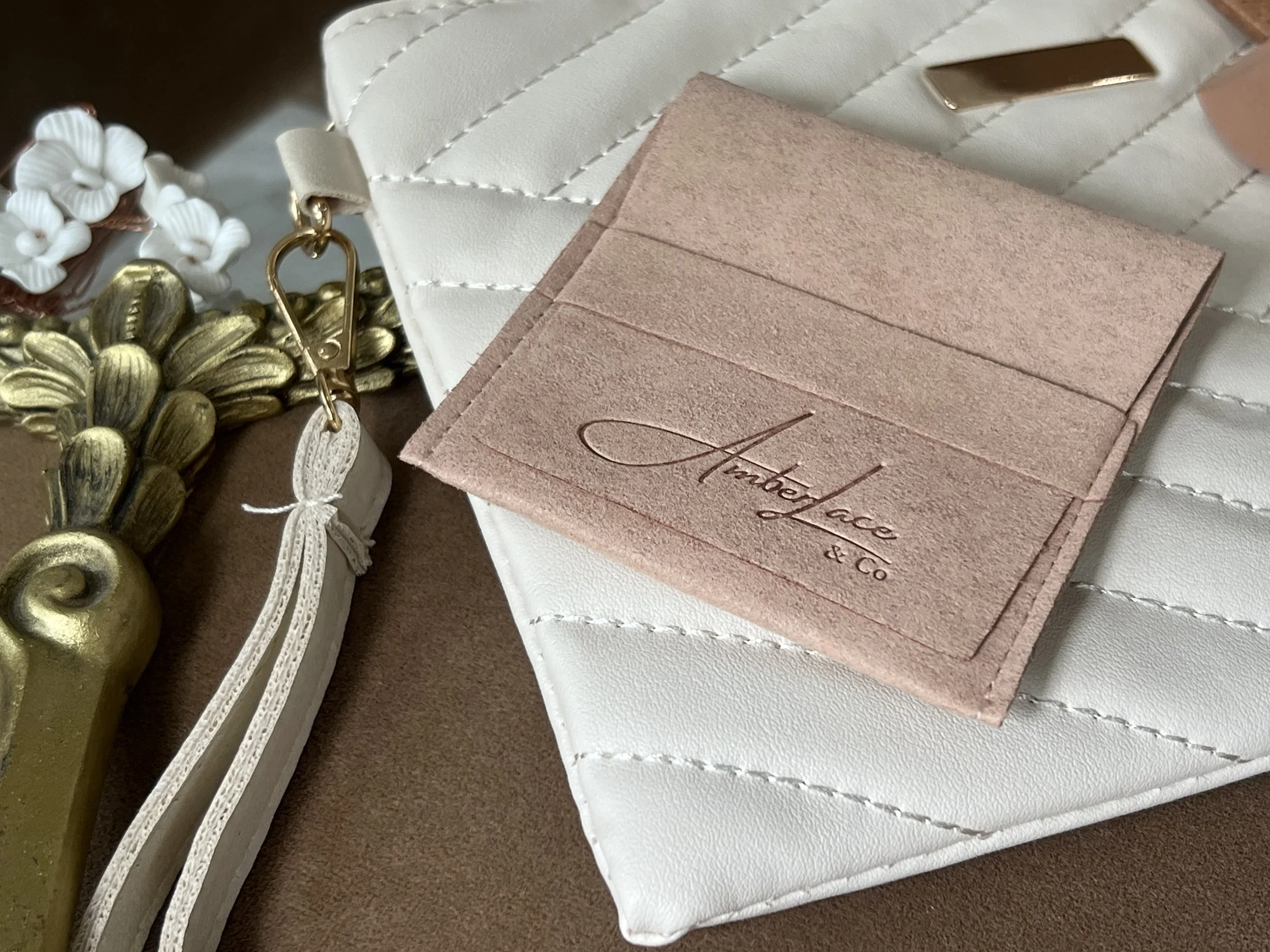 Close-up of a white quilted purse with a gold metal accent, a pink suede cardholder with the brand name 'Amberlace & Co.' embossed on it, and a white flower-shaped keychain with gold accents.
