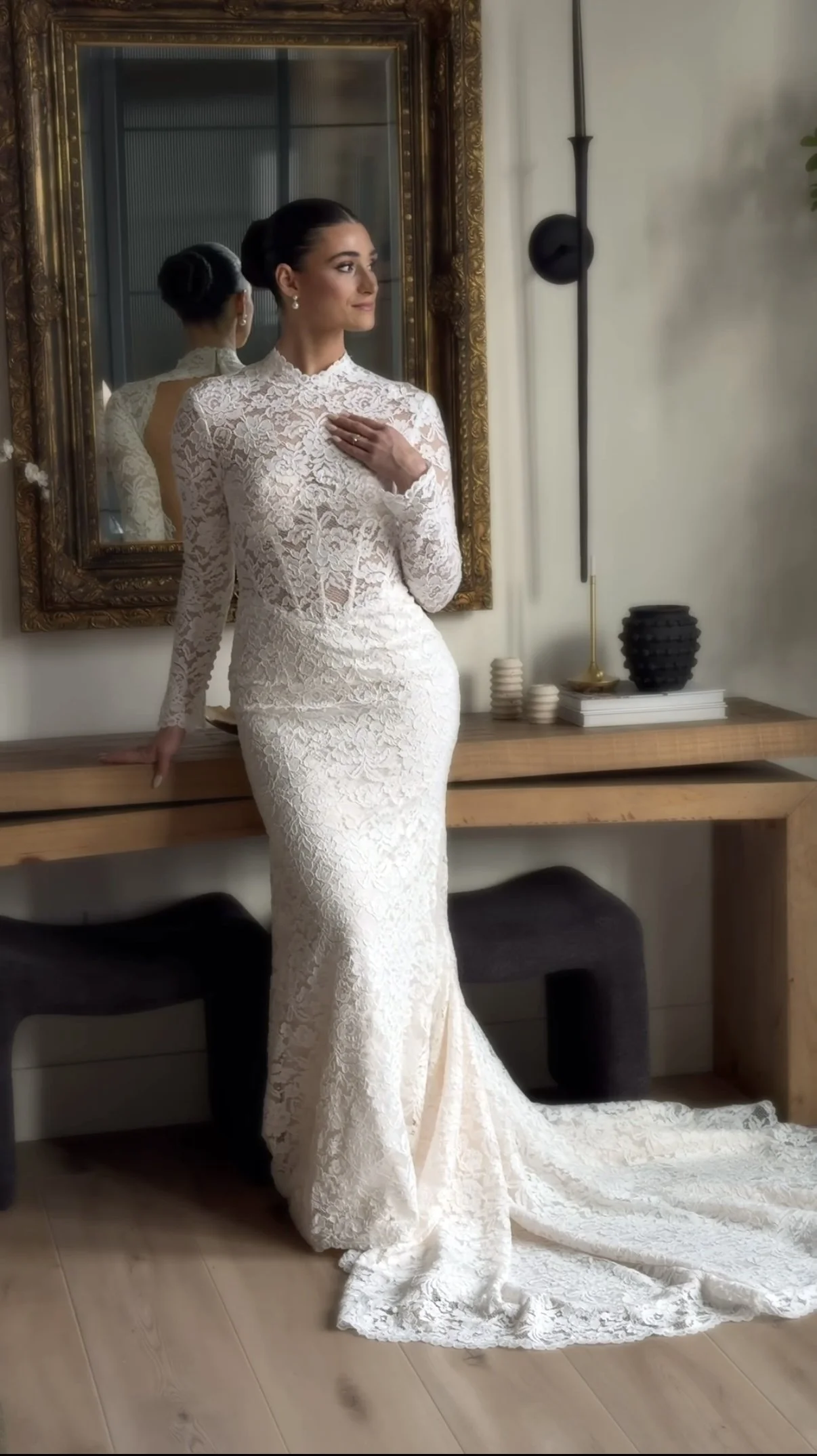 A woman in a white lace wedding dress posed indoors near a mirror and a wooden sideboard with decorative objects.