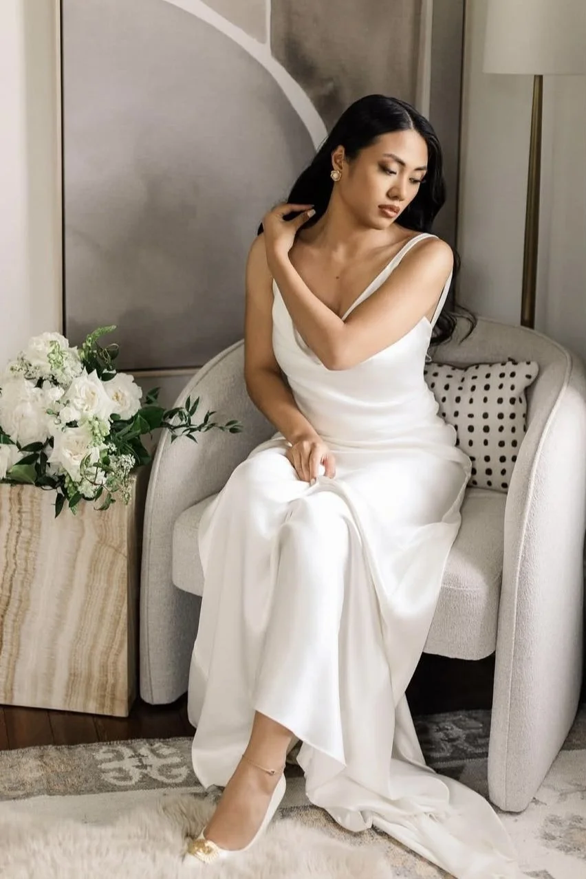 A woman in a white dress sitting on a beige sofa, touching her hair, next to a bouquet of white flowers and a pillow with black polka dots.