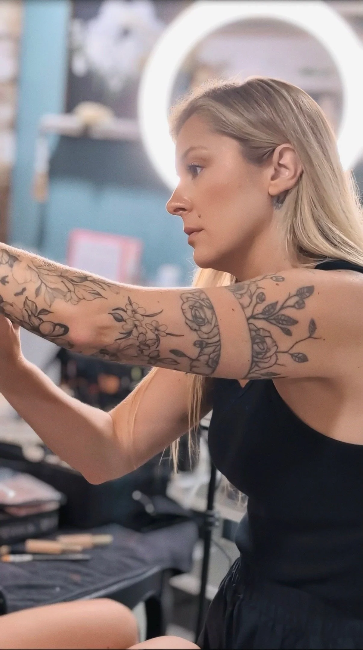 A woman with long blonde hair and floral tattoos on her arm, seated at a makeup station, applying makeup with a focused expression in a brightly lit room.