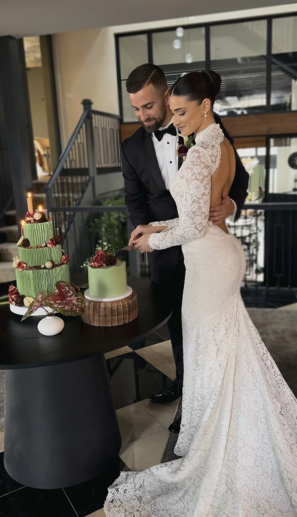 A bride and groom cutting their wedding cake indoors, with two cakes on the table, one decorated with strawberries and candles, and the other with a smooth green frosting.