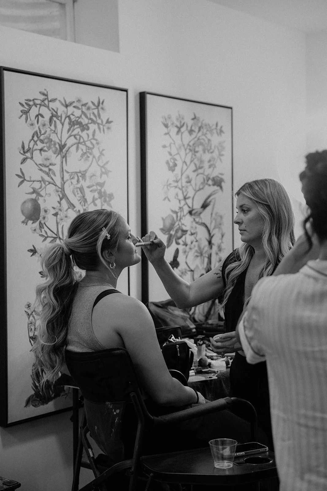 A woman sitting in a chair getting her makeup done by a makeup artist in a room with nature-themed artwork on the wall.
