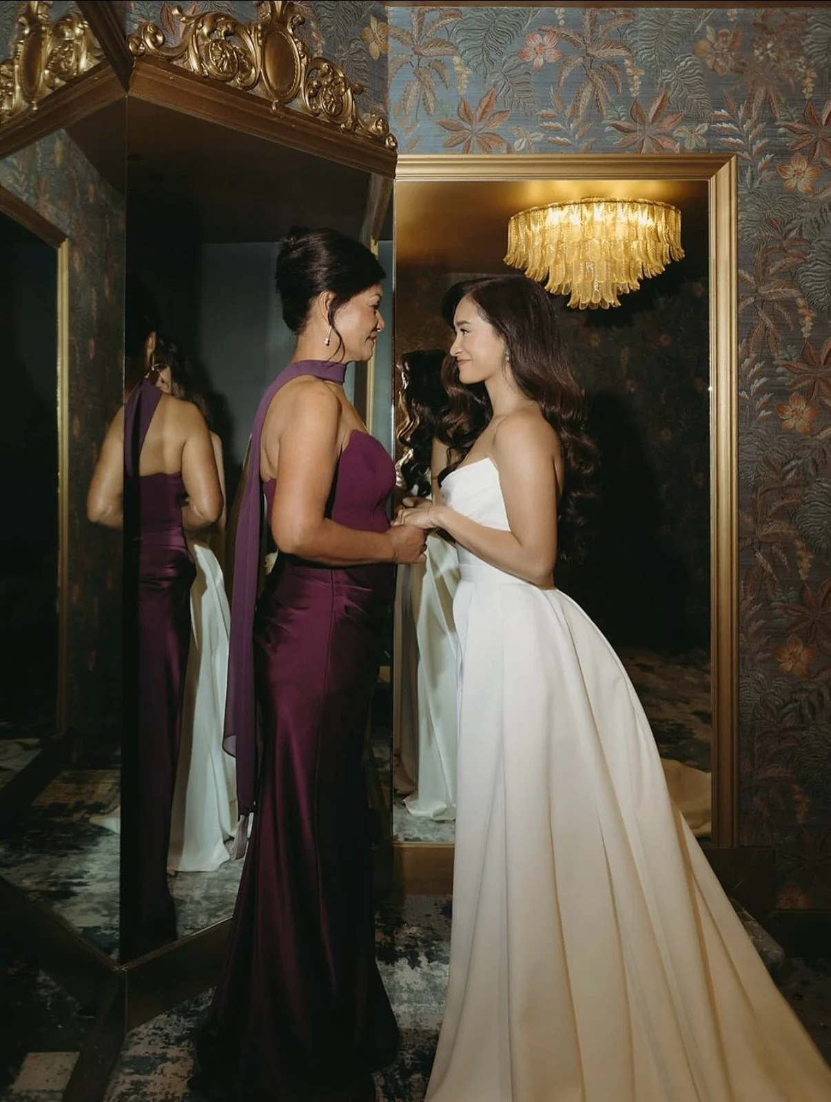 A woman in a purple gown and a woman in a white gown holding hands in front of a mirror, in an elegant room with floral wallpaper and a chandelier.