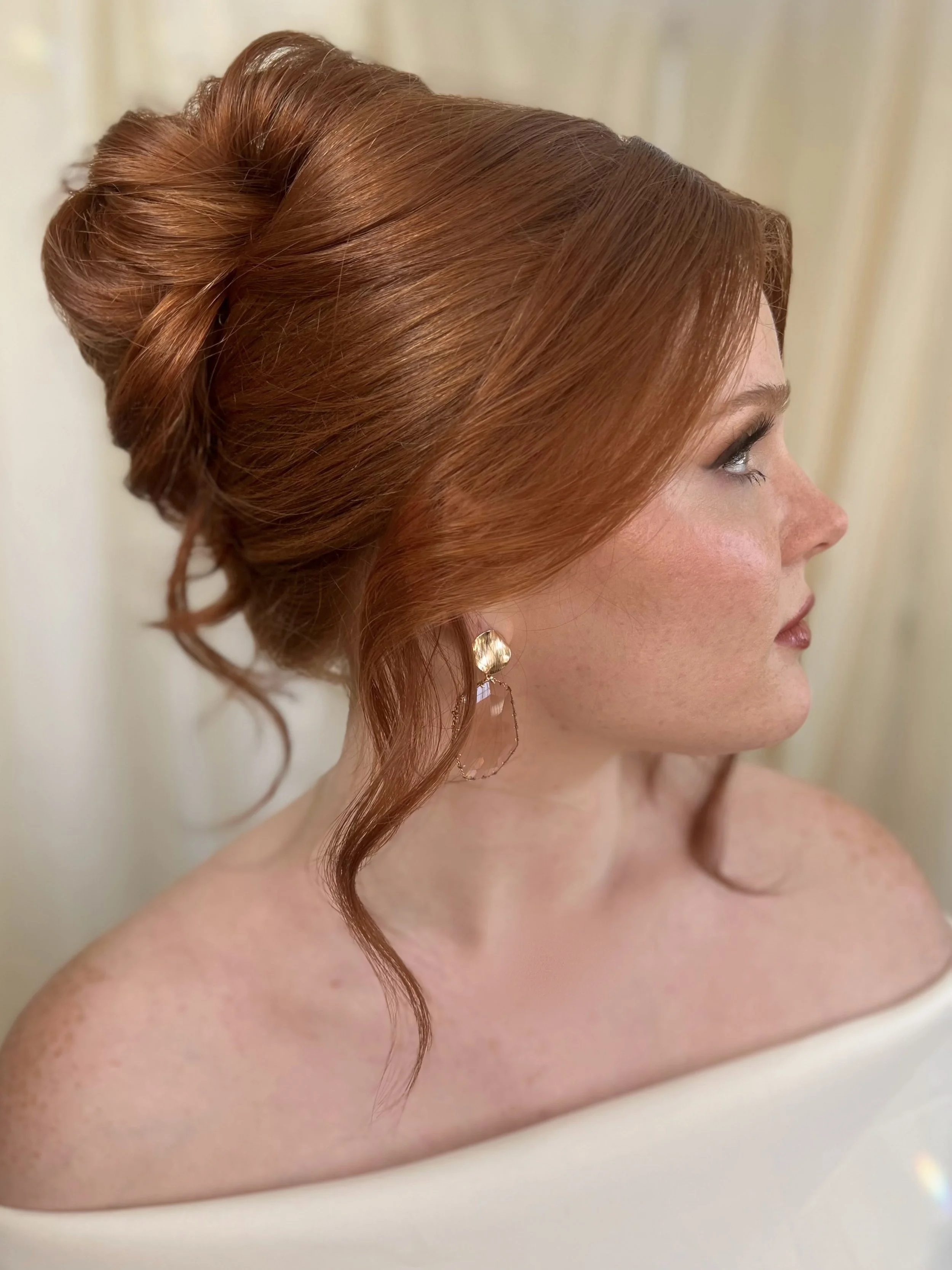 A woman with red hair styled in an elegant updo, wearing a gold and clear earring, and a white off-shoulder top, looking to the side with a neutral expression.
