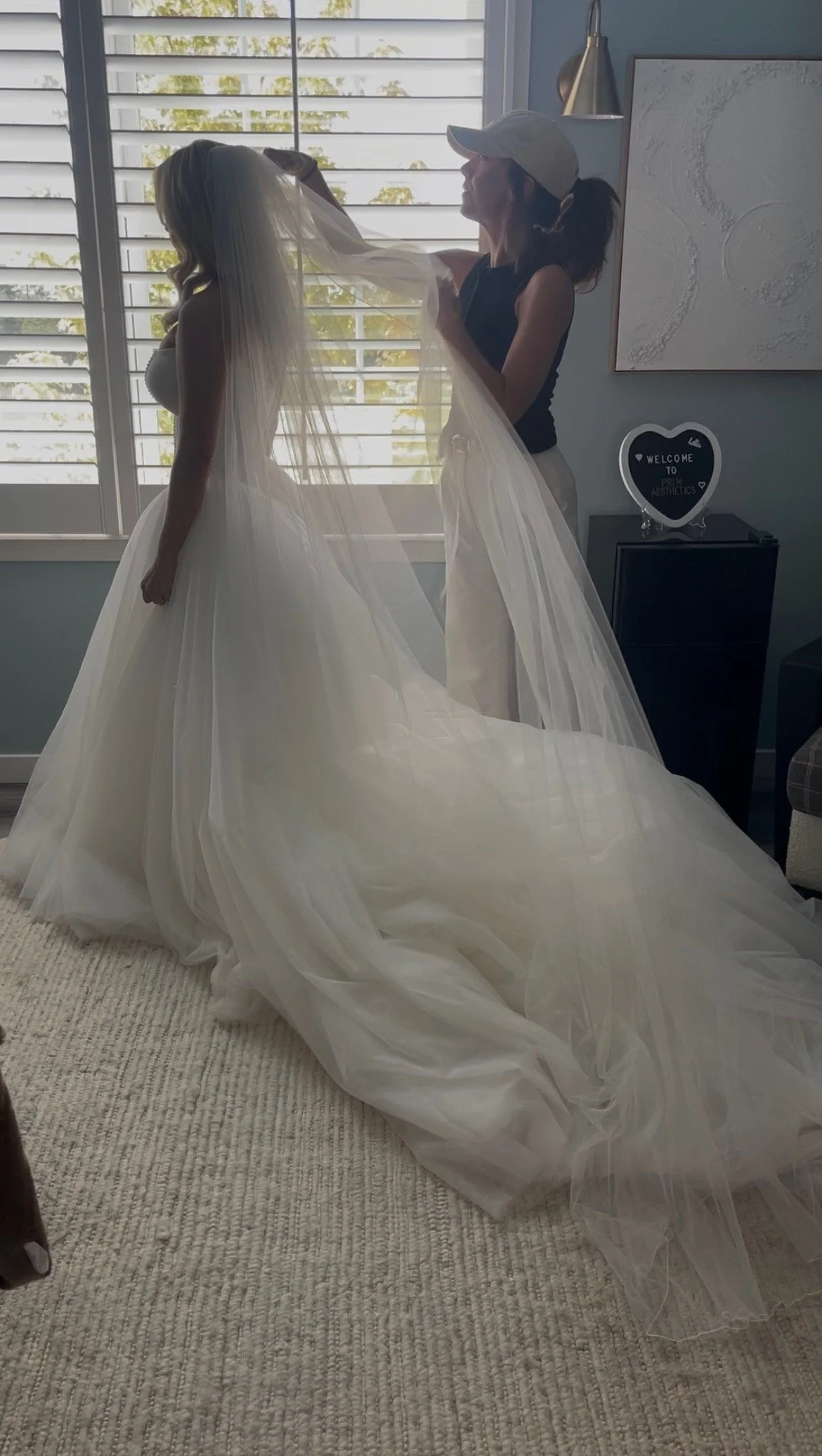 A bride is getting ready with assistance from a woman, possibly a stylist, in a room with light blue walls, white window blinds, and modern decor. The bride is wearing a voluminous, elegant white wedding gown and veil.