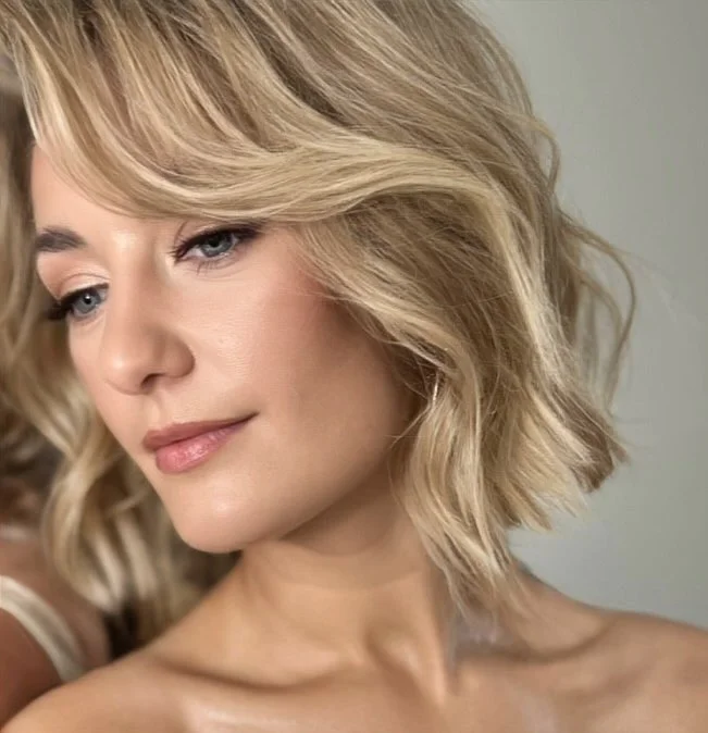 Close-up of a blonde woman with wavy hair looking down with a soft expression, wearing natural makeup.