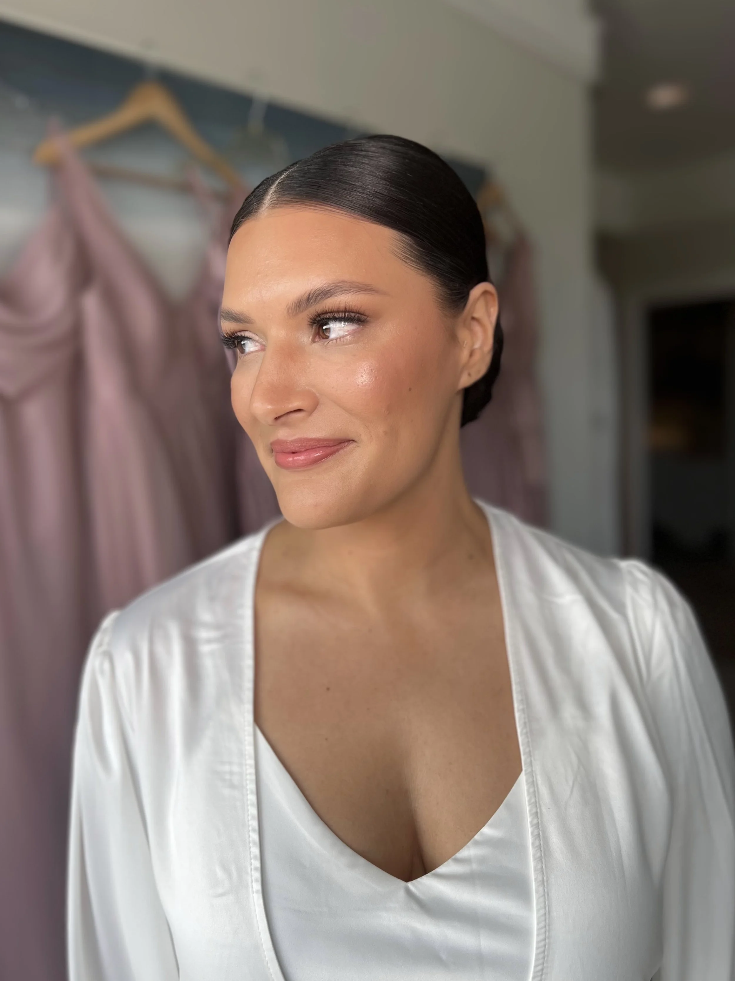 A woman with dark hair pulled back, wearing a satin white robe, standing in a room with pink dresses hanging on a rack behind her.