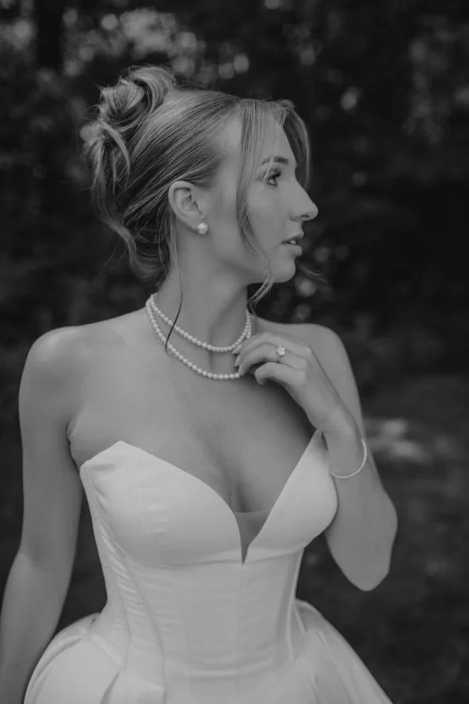 A woman in a strapless wedding dress with pearl jewelry, styled hair, and earrings, standing outdoors with trees in the background.