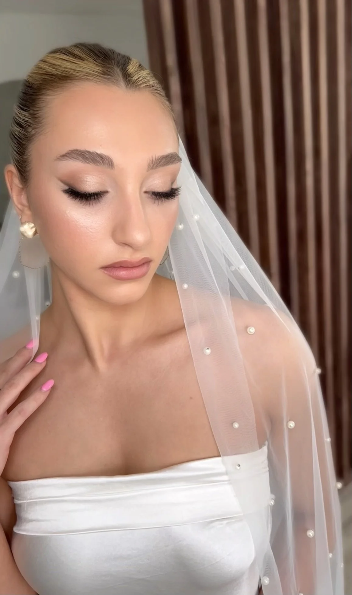 A bridal portrait of a woman with blonde hair pulled back, wearing pearl earrings and a silky white strapless dress, with a sheer veil decorated with pearls draped over her shoulder, and soft makeup with nude lipstick and subtle eye makeup.