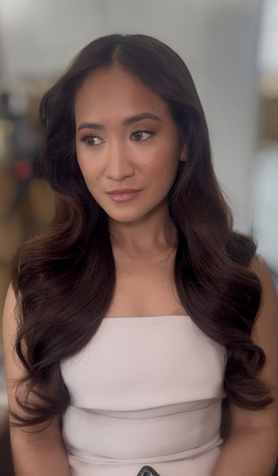 A young woman with long, dark wavy hair and light makeup, wearing a sleeveless white dress, is looking slightly off to the side in an indoor setting.