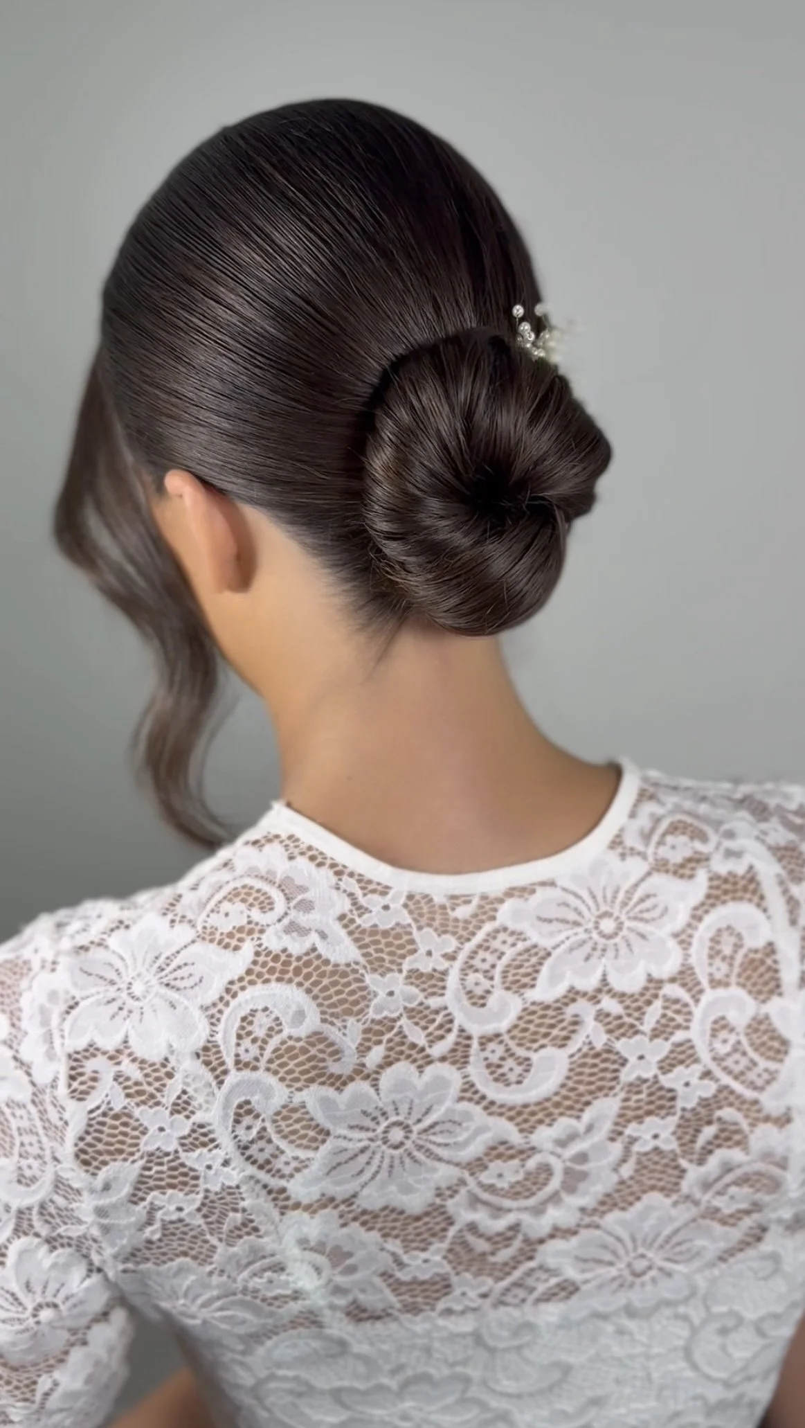 Back view of a woman with a sleek, elegant low bun hairstyle adorned with small decorative pearls, wearing a white lace top with floral patterns.
