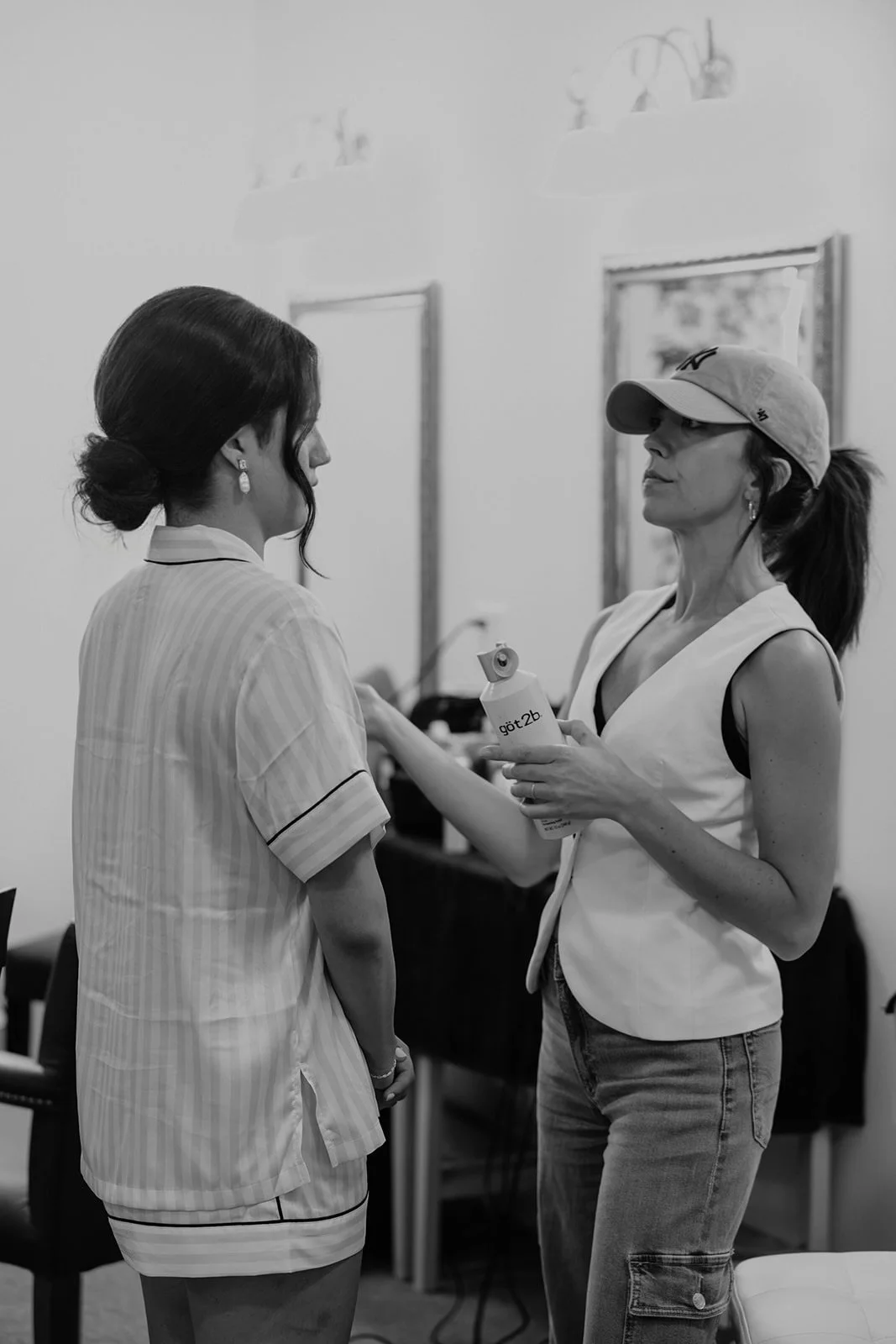 Two women engaged in conversation indoors, with one holding a water bottle, the other dressed in striped pajamas. The woman holding the water bottle is wearing a cap and sleeveless top, while the other has short hair styled in a bun.