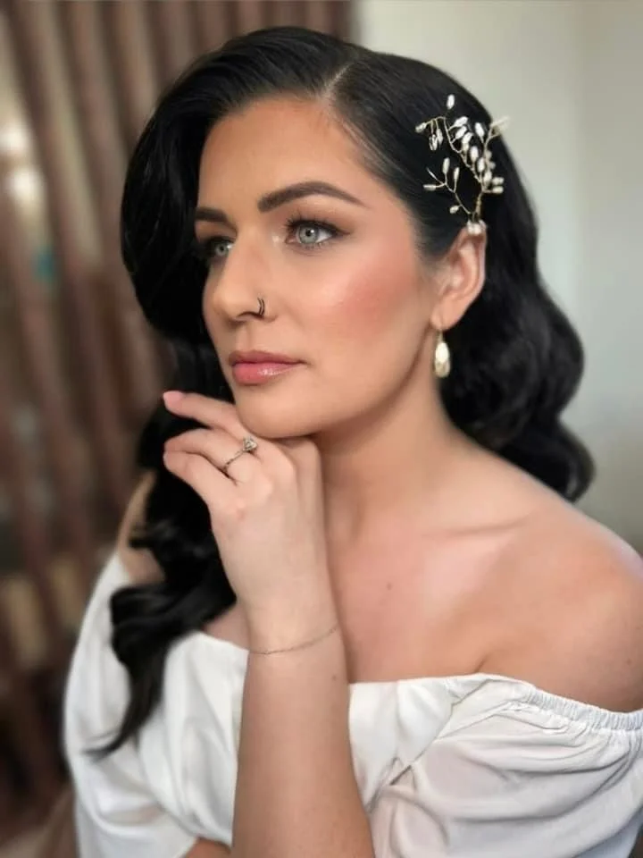 A woman with dark wavy hair, wearing a white off-the-shoulder top, gold earrings, and a decorative hairpin, resting her chin on her hand.
