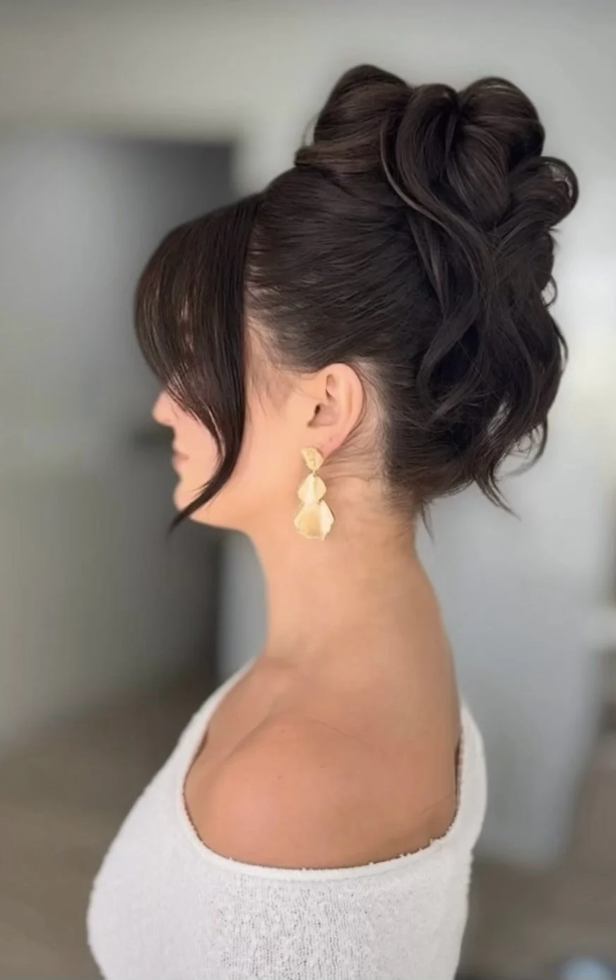 Woman with dark brown, styled hair in an updo with curls, wearing large gold earrings and a white off-the-shoulder top.