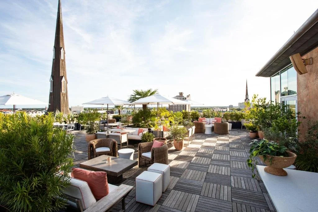 Rooftop garden with outdoor seating, umbrellas, and potted plants, with city skyline and church spires in the background.