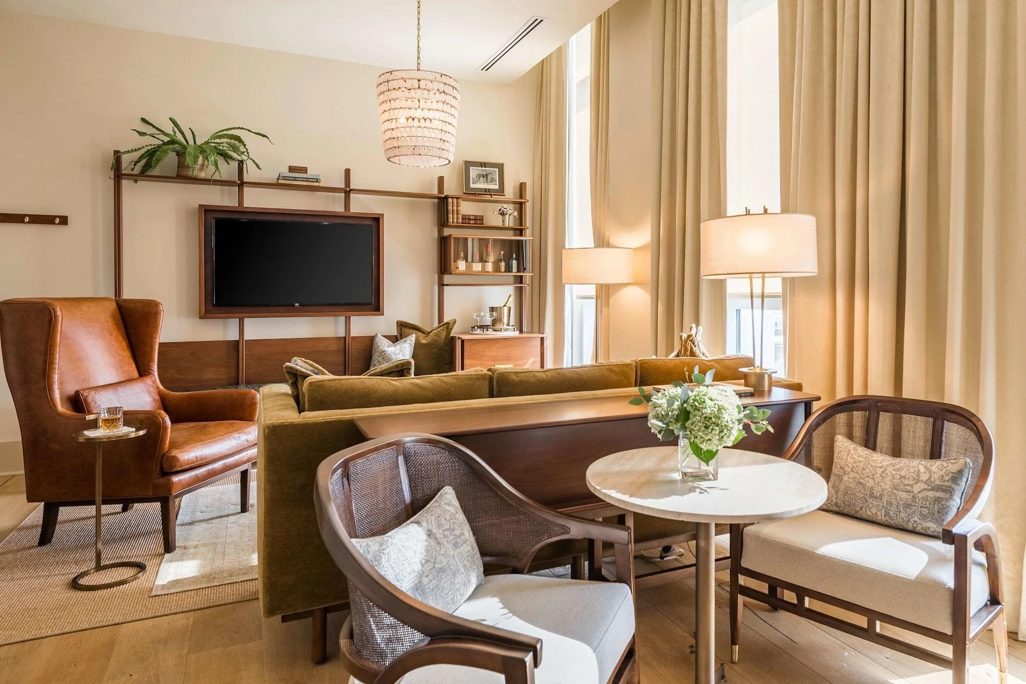 Living room with a brown leather armchair, green sofa, round coffee table with a flower arrangement, two armchairs, a TV mounted on a wall, and beige curtains.