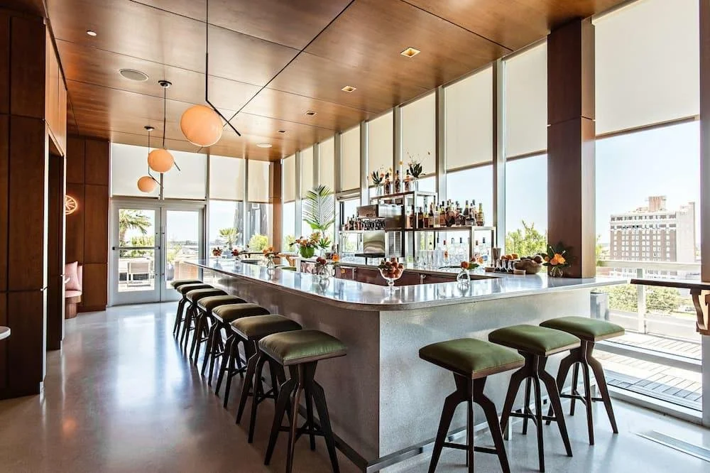 Interior of a modern bar or restaurant with a marble counter, bar stools with green seats, pendant lights, large windows, and a city view.