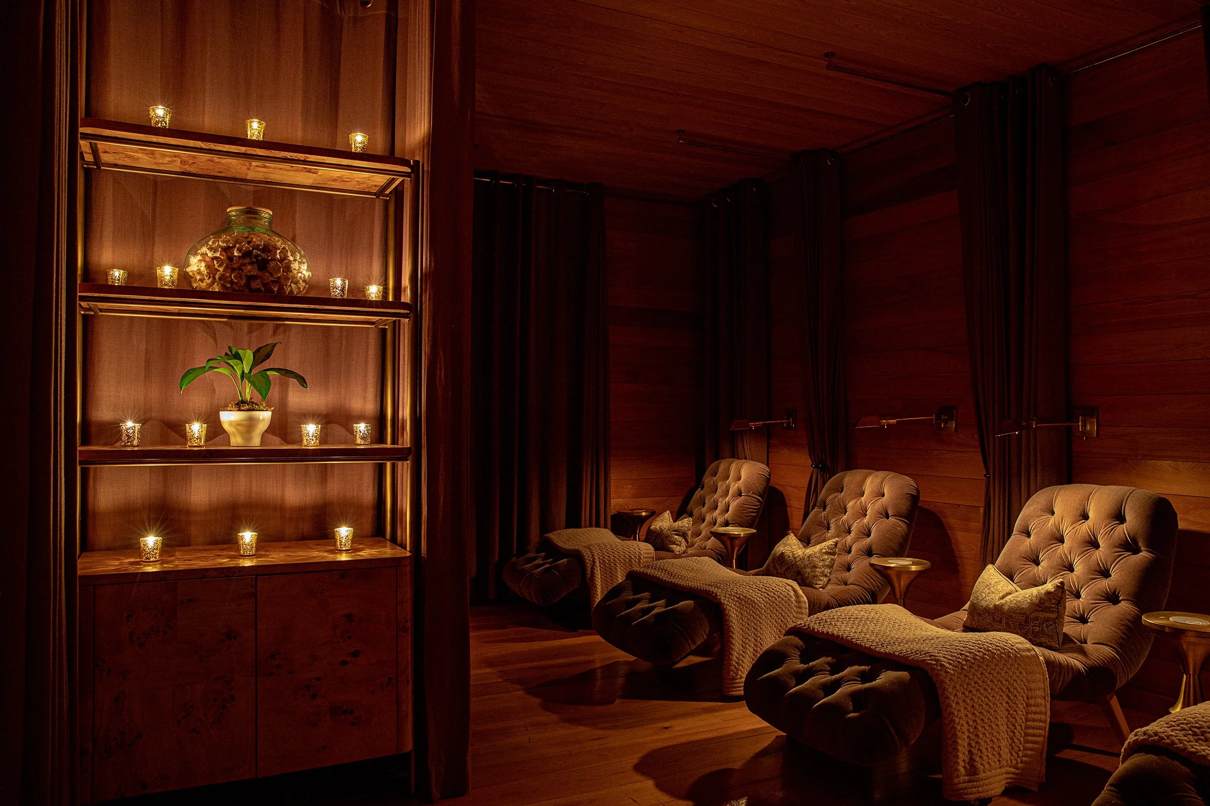 A cozy room with three plush recliner chairs, wooden walls, curtains, and small tables, illuminated by candlelight and soft lighting, with a shelving unit containing a potted plant, candles, and decorative items.