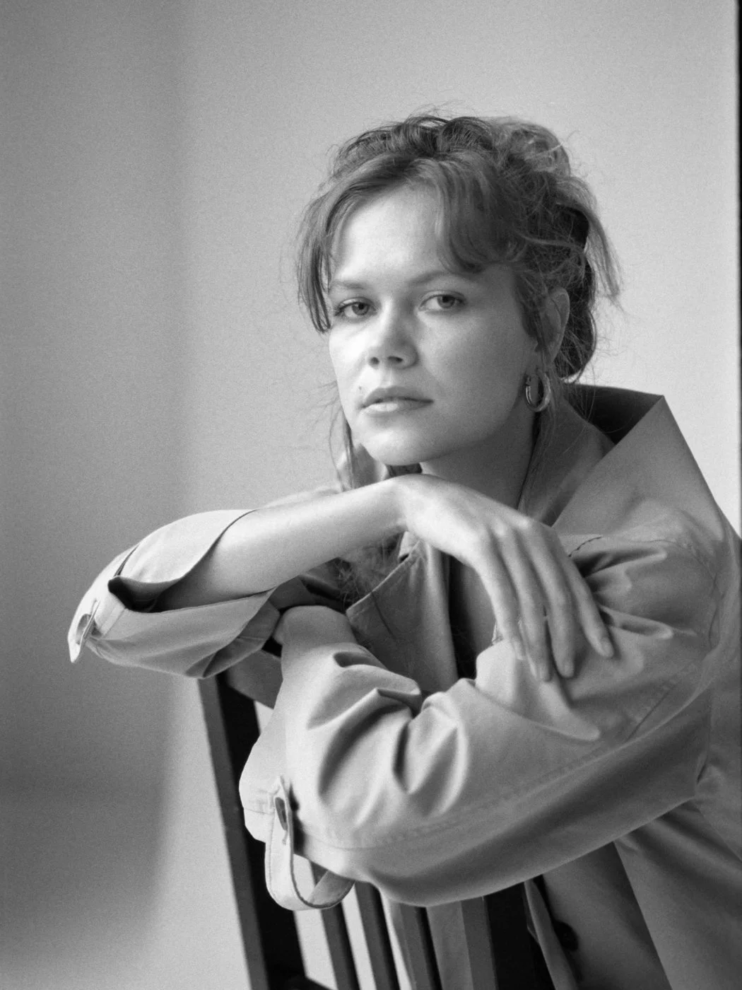 Black and white portrait of a woman with wavy hair, wearing earrings and a jacket, sitting with her chin resting on her folded arms, looking at the camera.