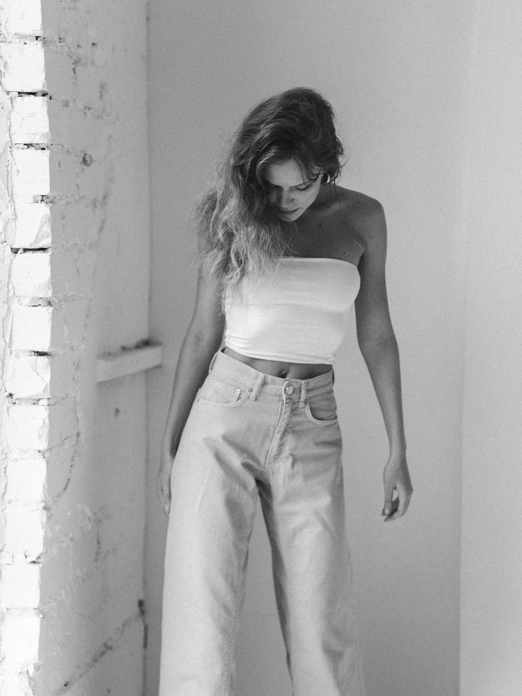 A woman with long, curly hair, dressed in a strapless white top and loose high-waisted pants, standing against a plain wall and a partially painted brick wall in black and white.