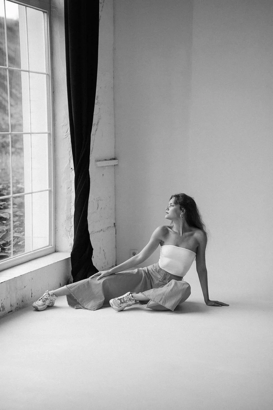 A woman sitting on the floor near a large window with curtains, looking out. The room has a simple, minimalist aesthetic with plain walls.
