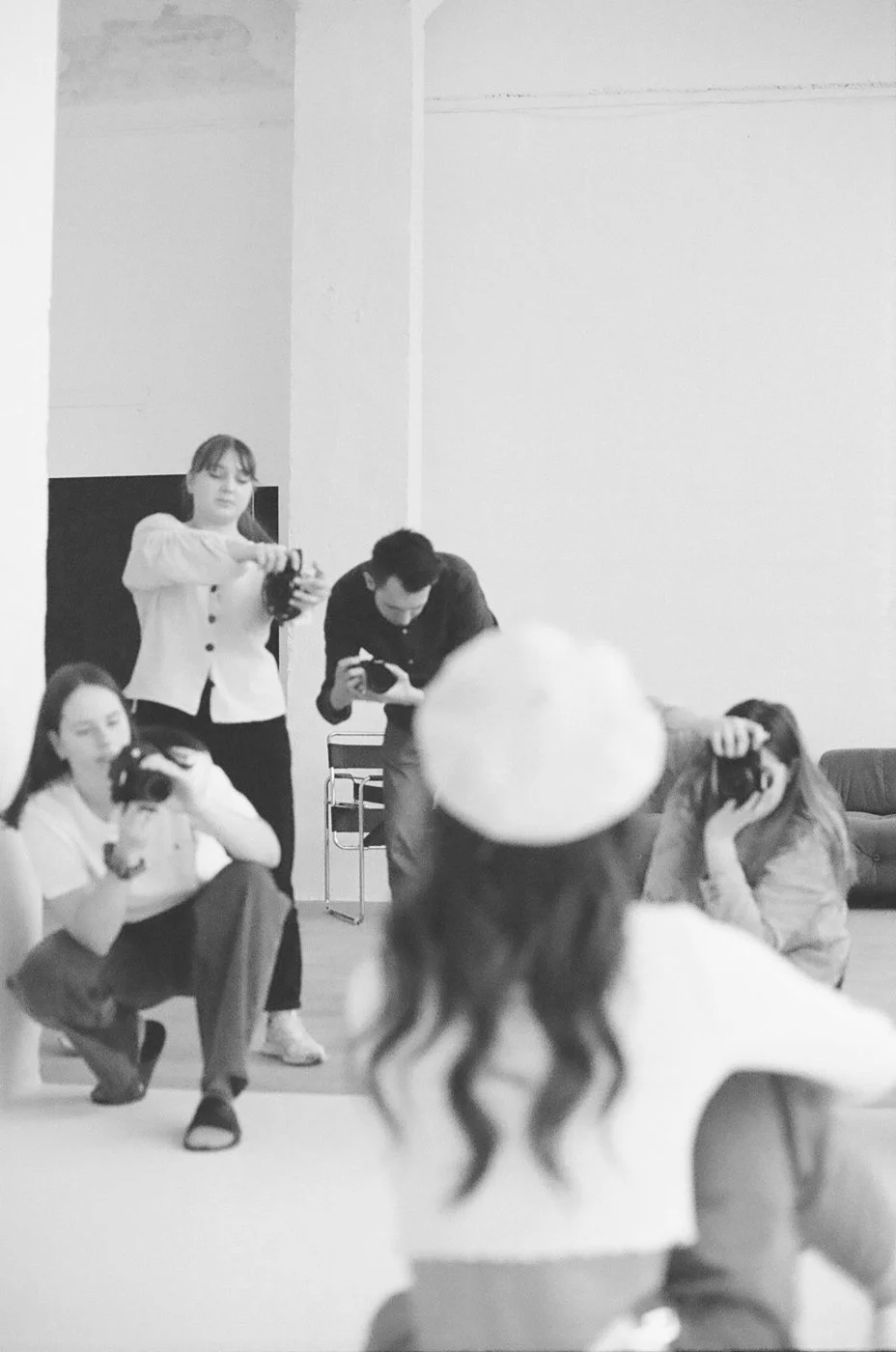 A black-and-white photo of a group of people in a room, some taking pictures with cameras. The photo is taken from behind a woman with long wavy hair and a beanie, capturing her from the back.