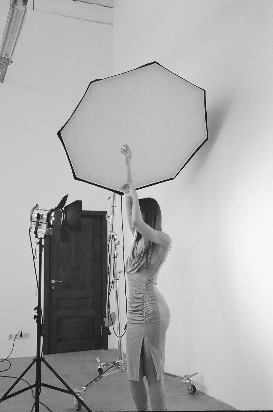 A woman in a fitted dress standing next to photography lighting equipment in a studio, reaching up towards a large octagonal softbox.