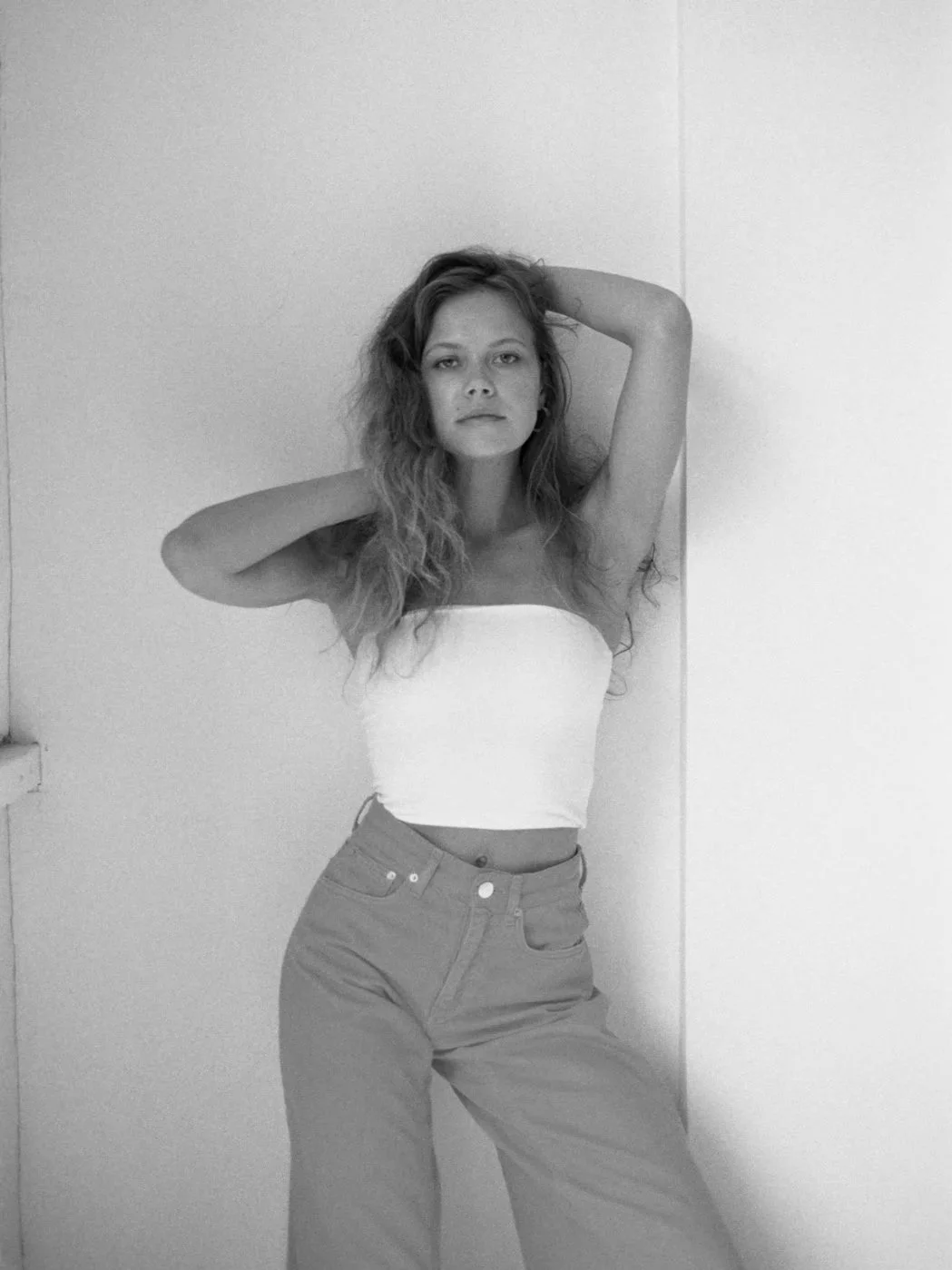 Black and white photo of a young woman with wavy hair, wearing a strapless white top and high-waisted pants, standing against a plain wall with one arm raised behind her head.