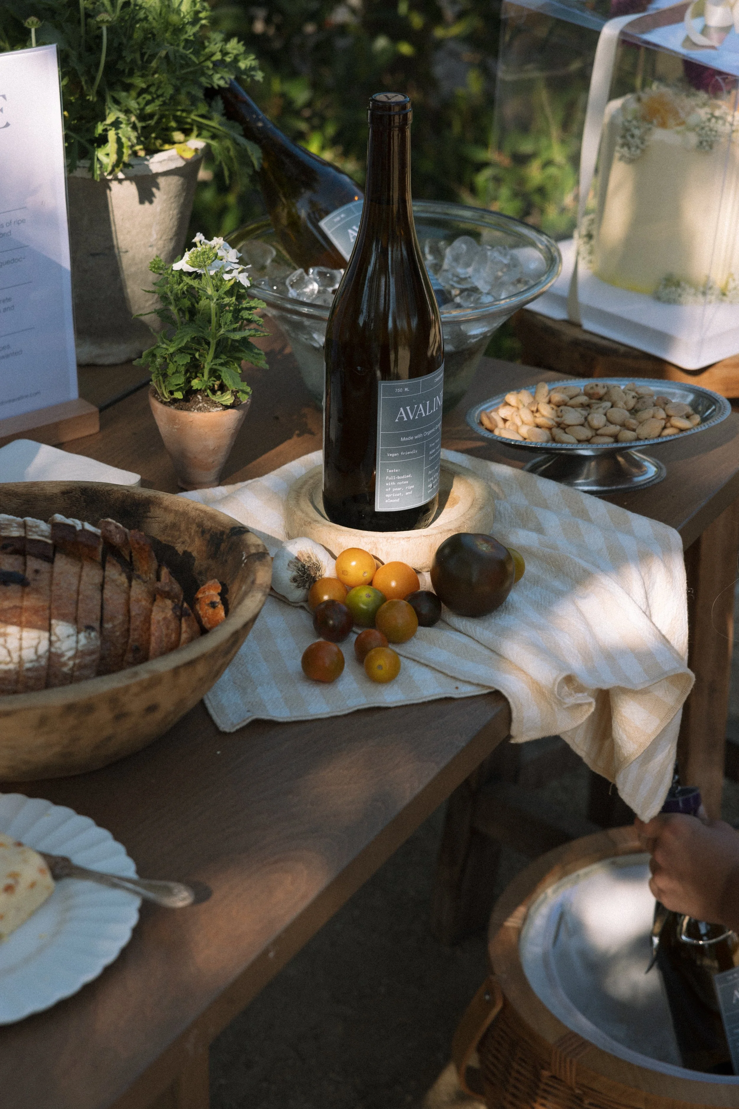 A table set with a wine bottle, grape tomatoes, a dark heirloom tomato, a loaf of sliced bread in a wooden bowl, a plate of flatbread, a bowl of nuts, a glass of ice water, and potted plants. The tablecloth is beige with white stripes.