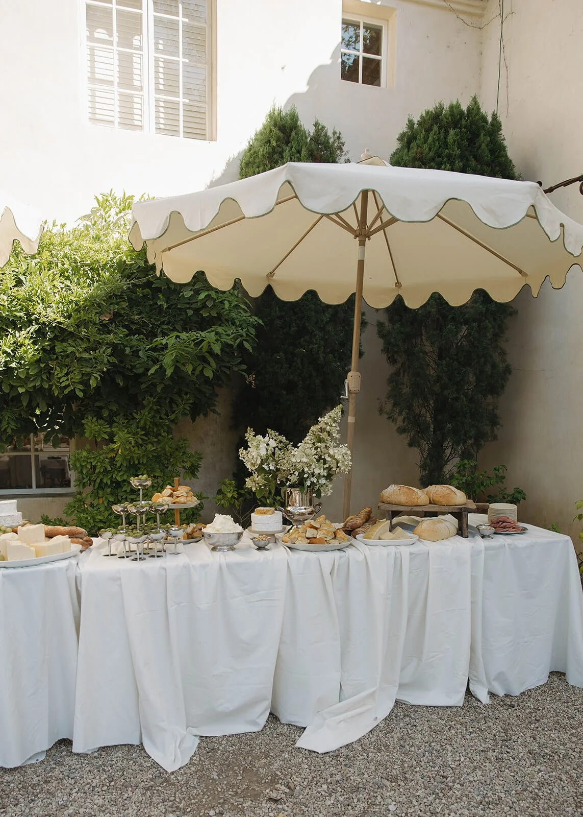 An outdoor buffet table covered with a white tablecloth, set under a large white patio umbrella. The table has a variety of food items including bread, cheese, and pastries, along with a bouquet of white flowers in a silver vase, surrounded by greenery and a white wall with a window.