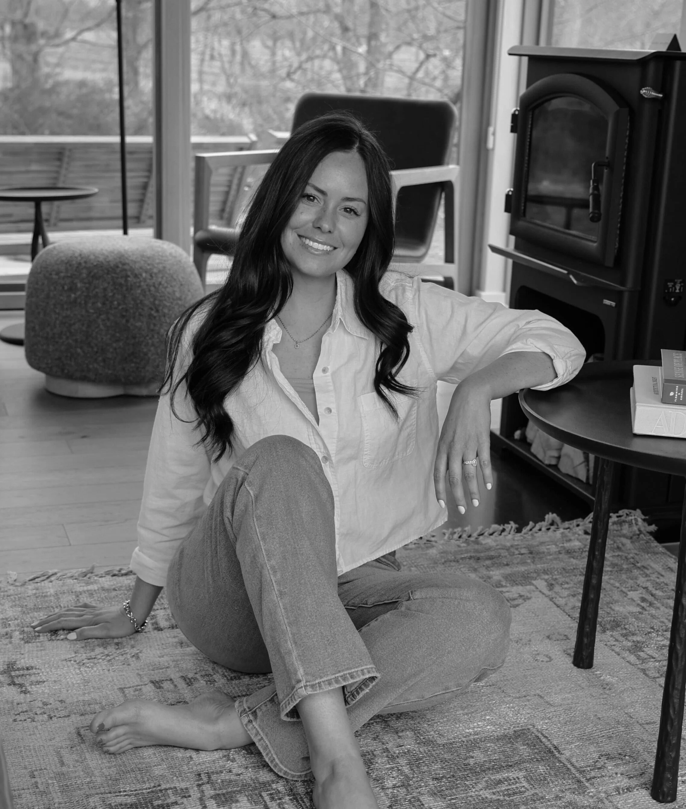A woman sitting on a rug in a living room, smiling at the camera, with a wood stove, a coffee table, and large windows in the background, black and white photo.