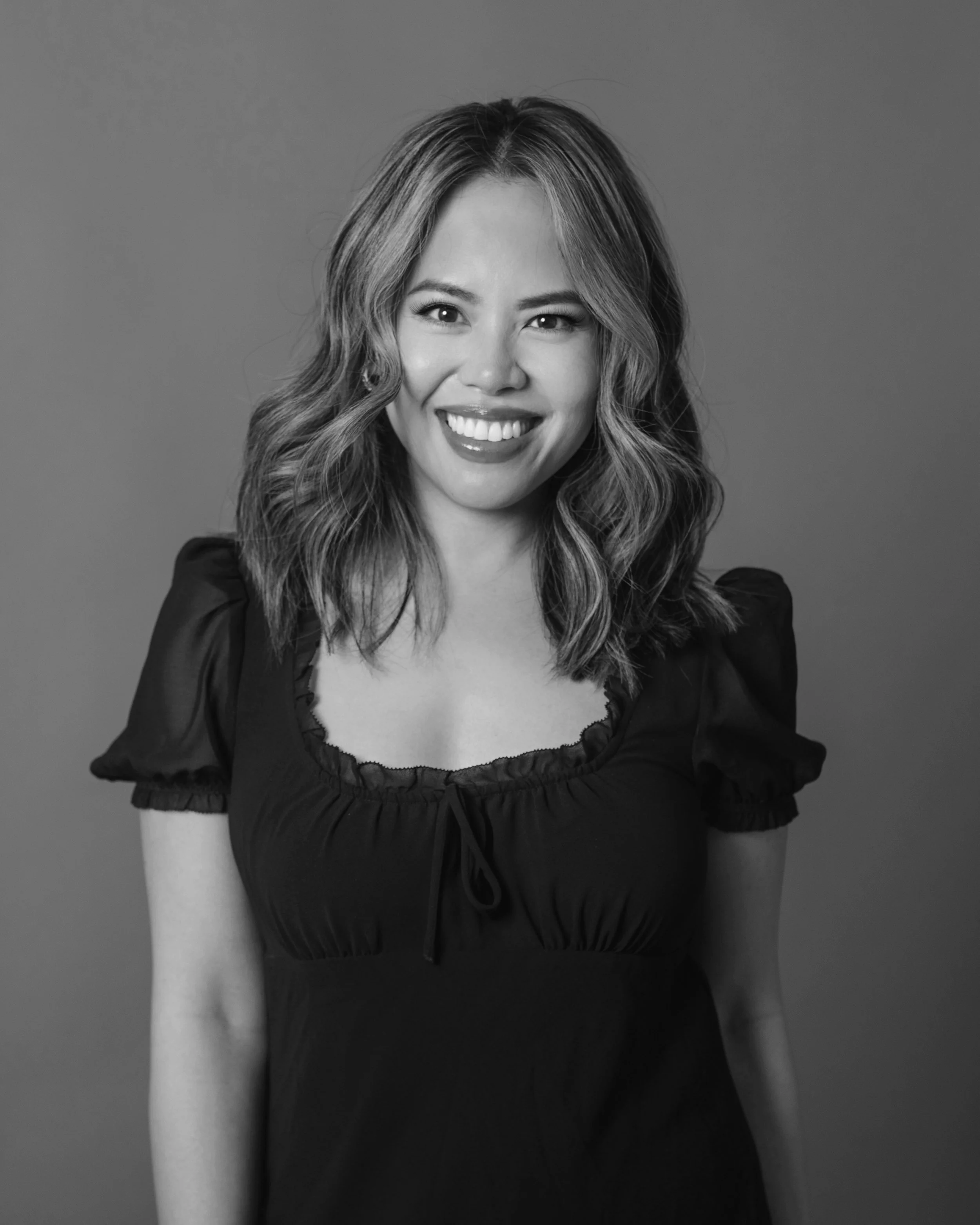 Black and white portrait of a smiling young woman with wavy shoulder-length hair, wearing a dark dress with puffed sleeves and a ruffled neckline.