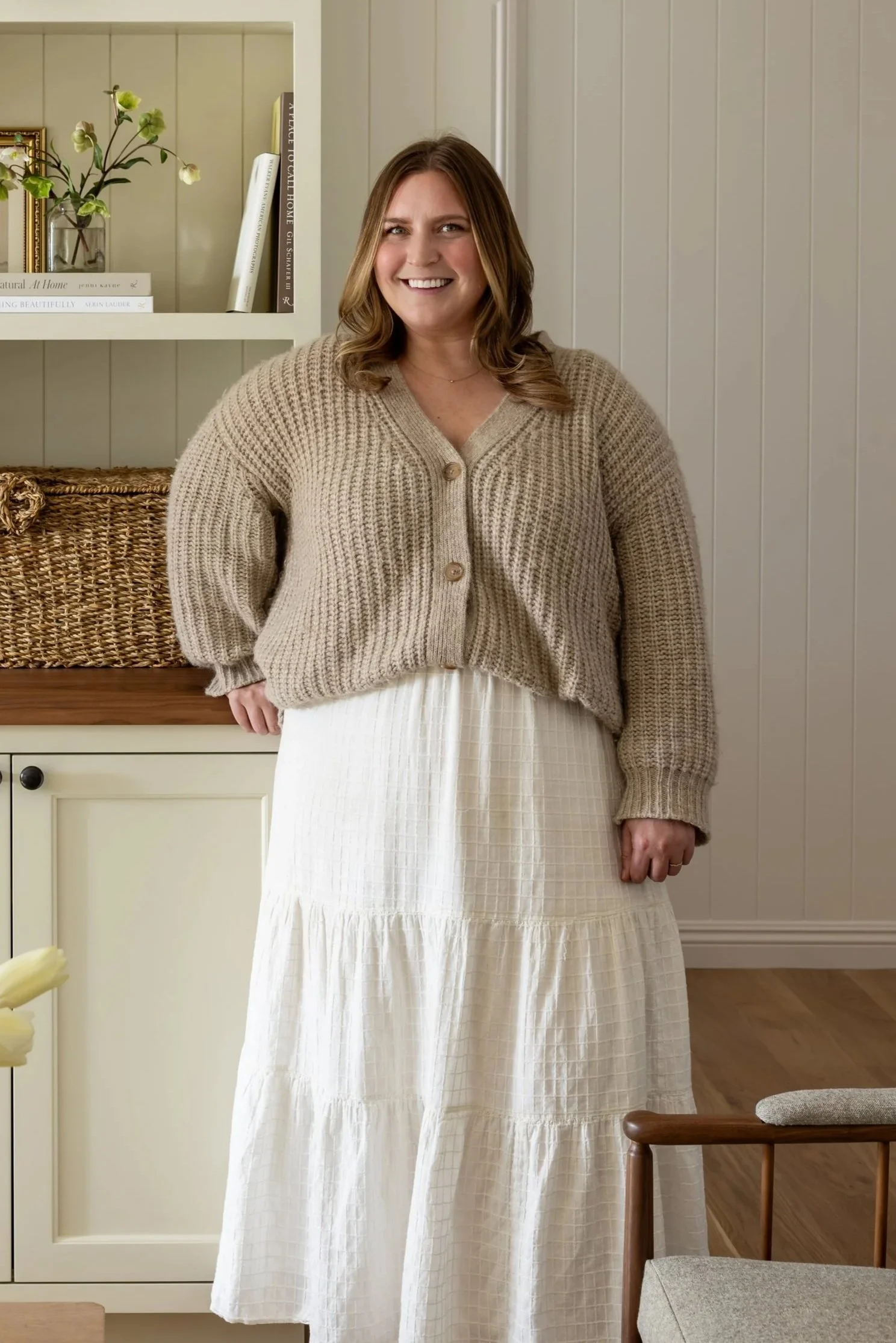 A smiling woman standing indoors in front of a white shelf with books and a vase, wearing a beige cardigan and a long white skirt.