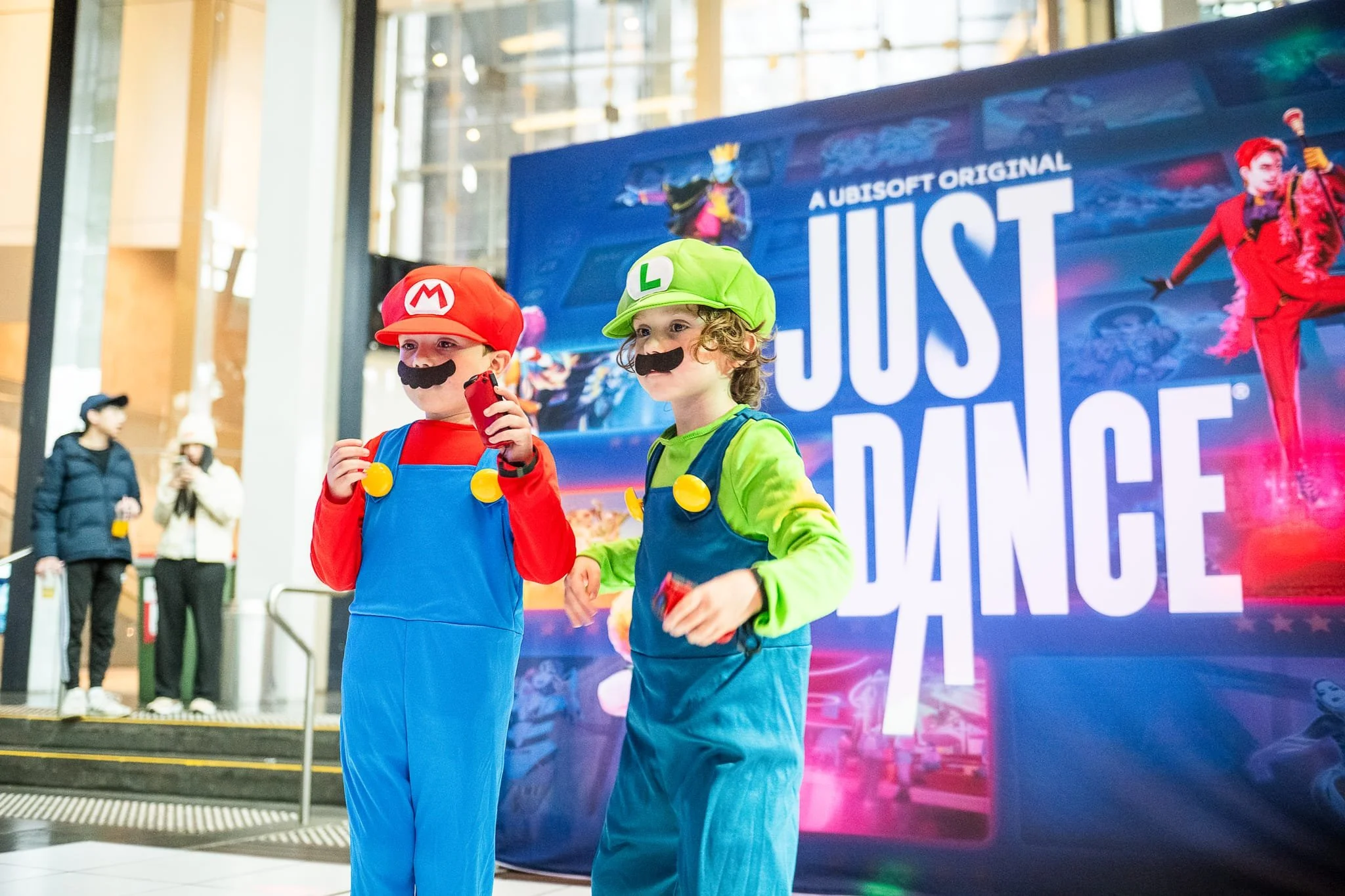 Two children dressed as Mario and Luigi from Super Mario, wearing costumes with hats, fake mustaches, and overalls, standing in front of a large movie poster for 'Just Dance' at a public event or mall.