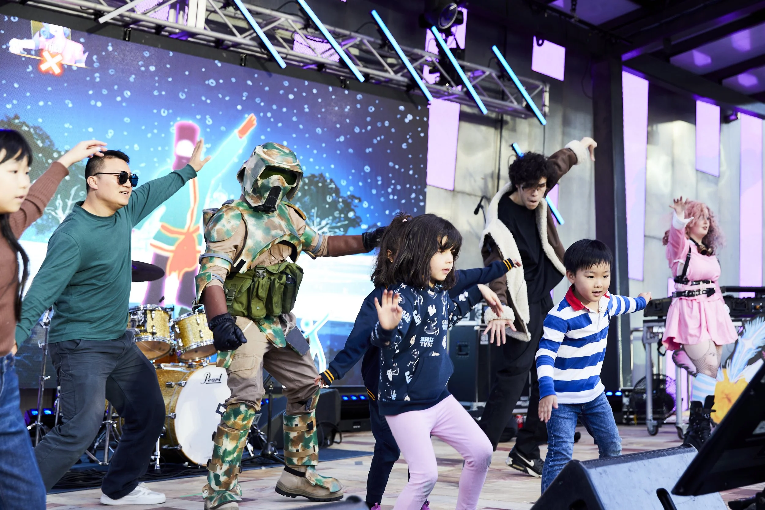 Group of children and adults dancing on stage with a colorful LED background, some wearing costumes including camouflage and pink clothing, with musical instruments in the background.