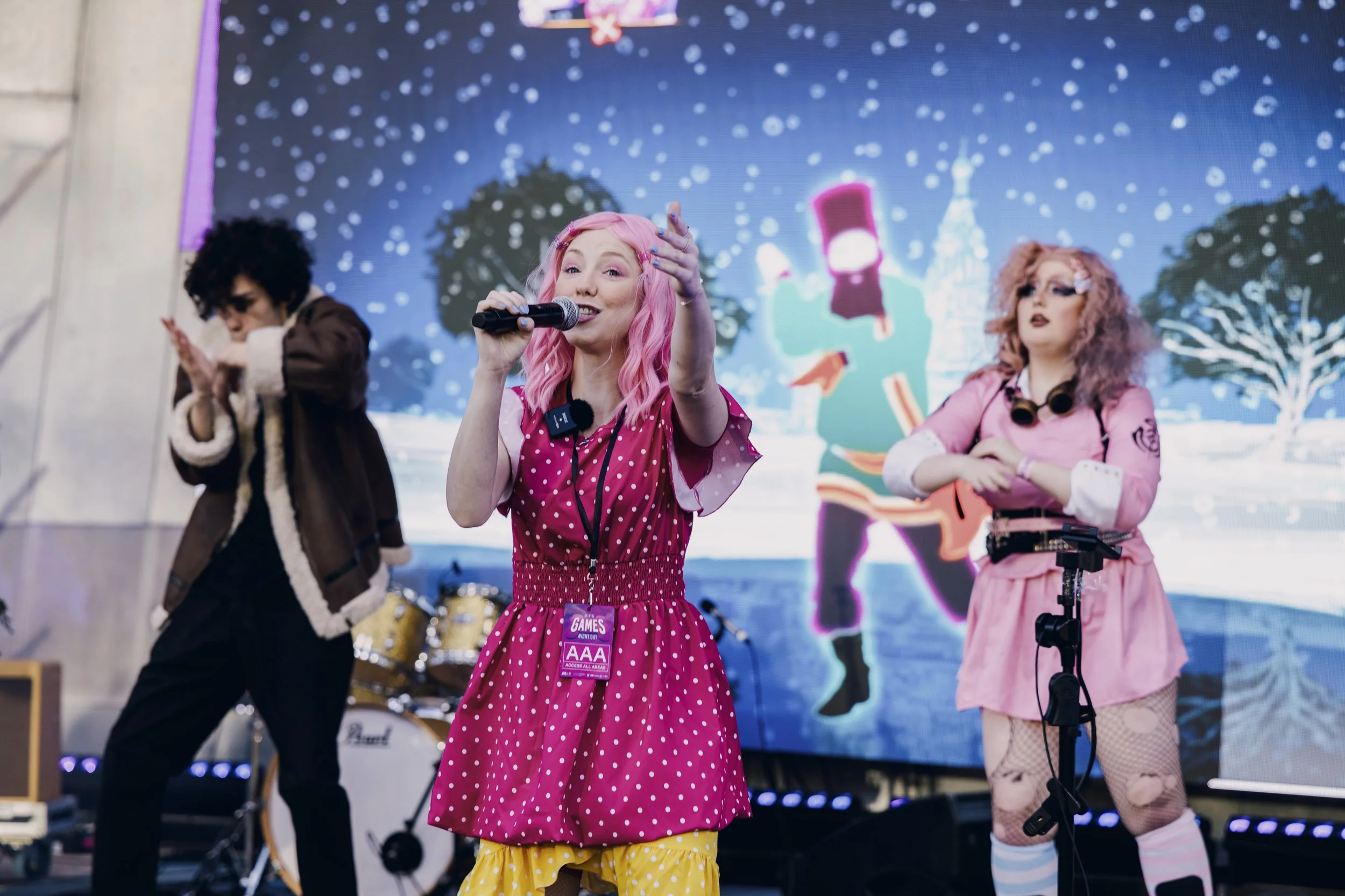 Three performers on stage during a holiday-themed event. The central woman with pink hair is singing into a microphone, wearing a pink polka dot dress with yellow ruffled shorts below. To her right, a woman with curly pink hair, glasses, and a pink s