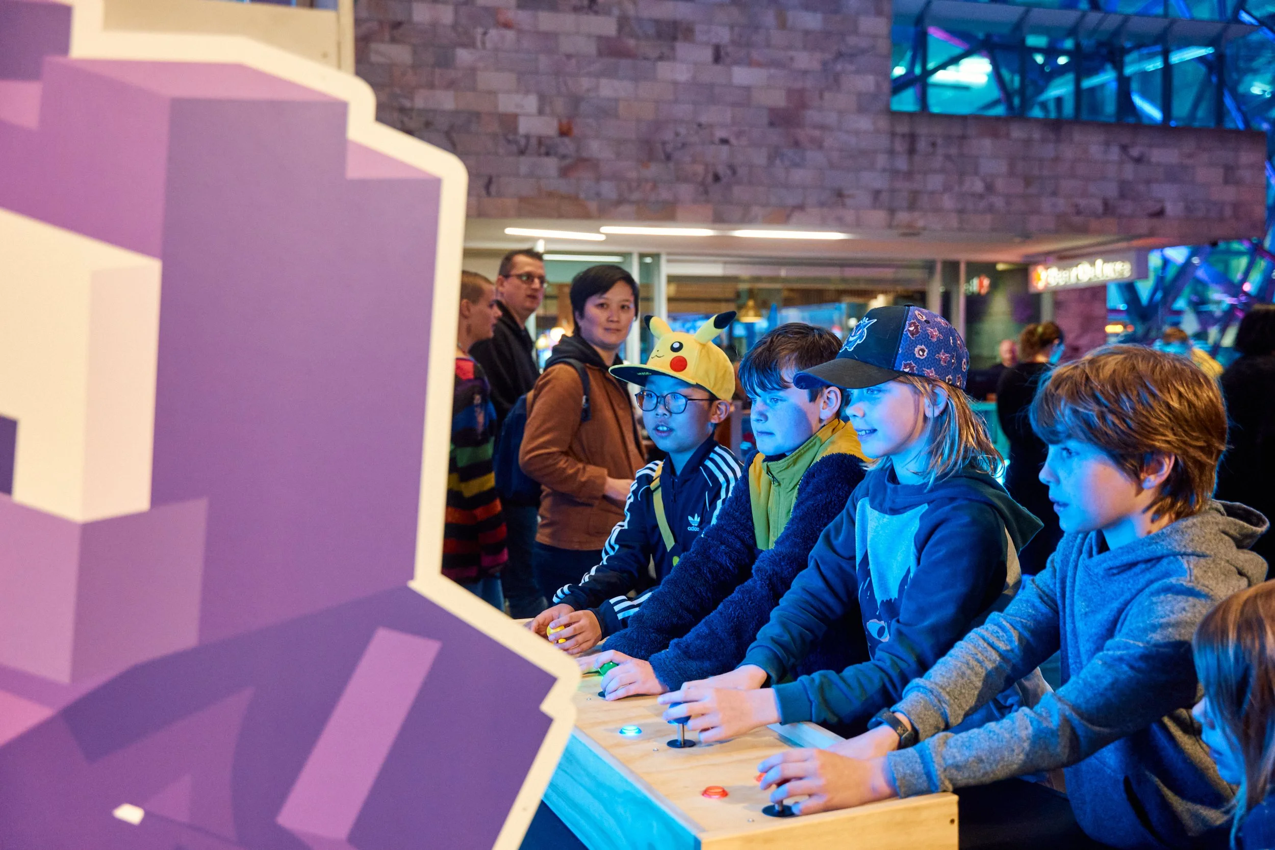 A group of children and teenagers playing an arcade game in a lively, colorful indoor space. Some are wearing hats and glasses, while others focus on the game.