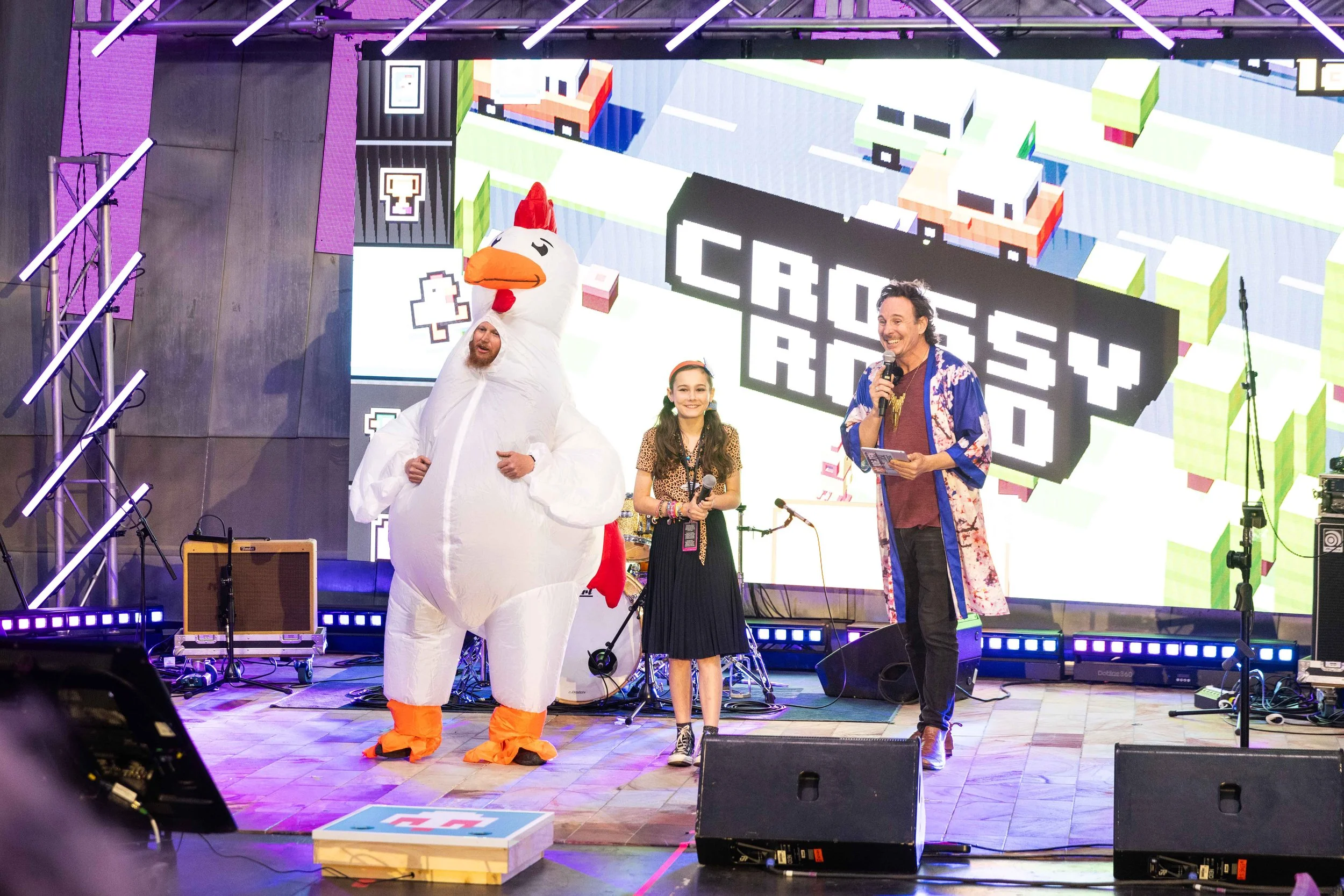 People on stage at an event, including a person dressed in a giant chicken costume, a young girl holding a microphone, and a man with a microphone, with a colorful digital background reading 'Crossy Road'.