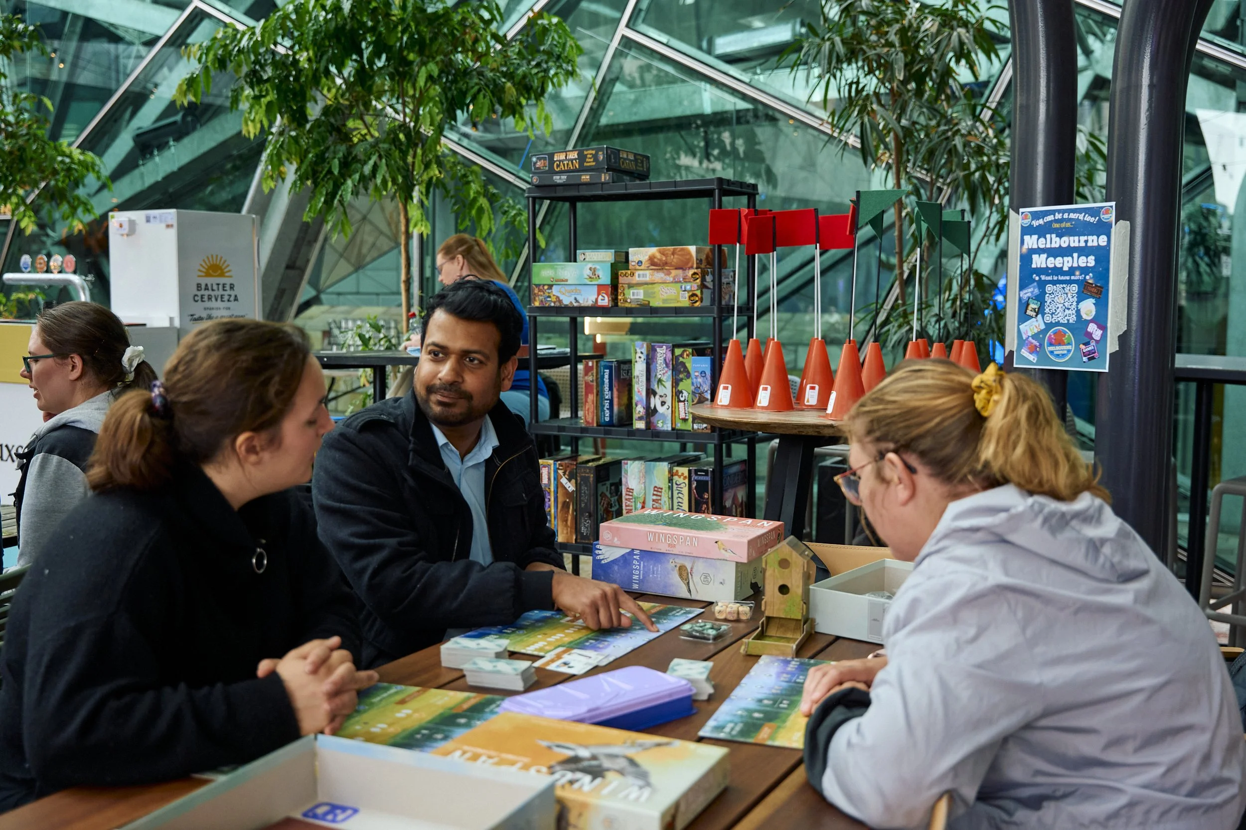 Group of four people playing board games at a table in an indoor space with glass architecture and greenery.