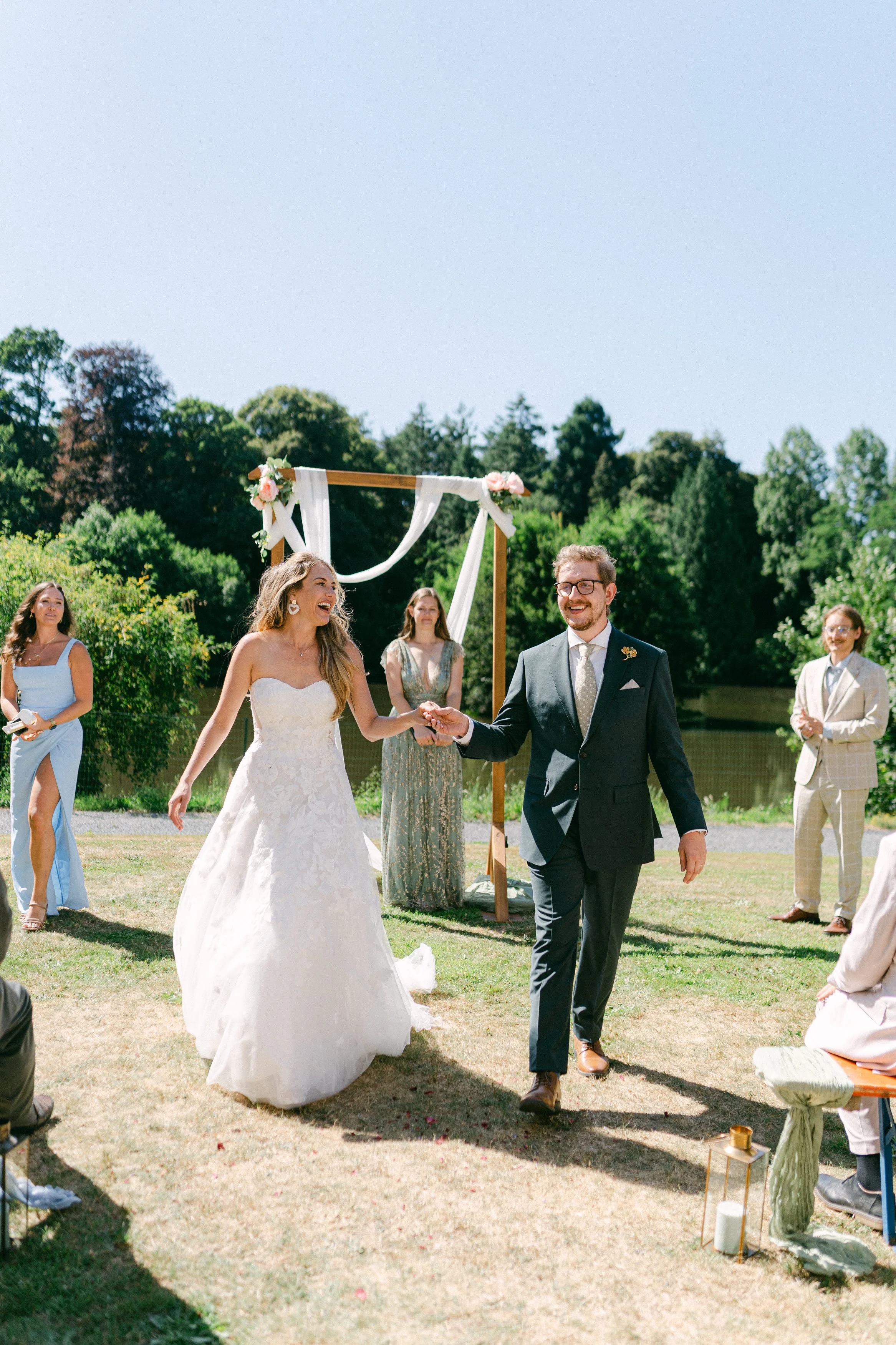 Un couple de mariés souriant lors d'une cérémonie en plein air, entouré de témoins, avec un décor floral et une scène en plein soleil.