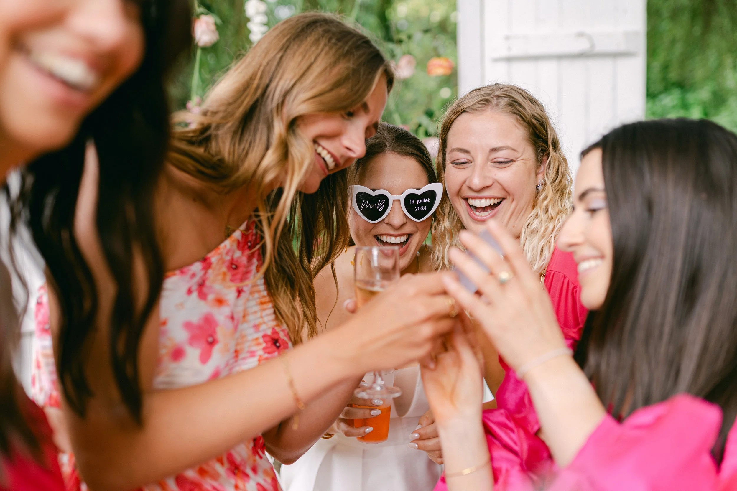 Groupe de femmes souriantes célébrant une occasion spéciale lors d'une fête, portant des vêtements colorés, avec des lunettes amusantes et tenant des verres de boisson, dans un environnement extérieur.
