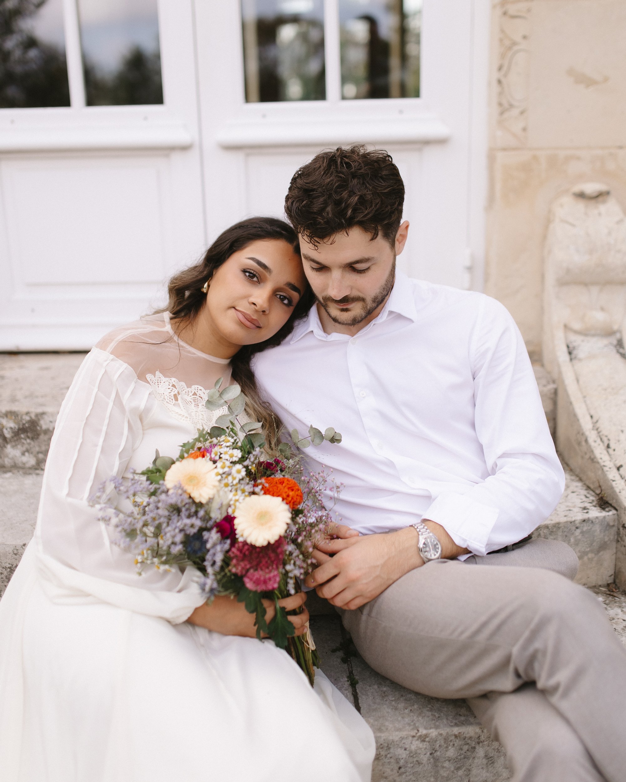 Un couple assis sur des marches, la femme tenant un bouquet de fleurs, ils se regardent avec tendresse.