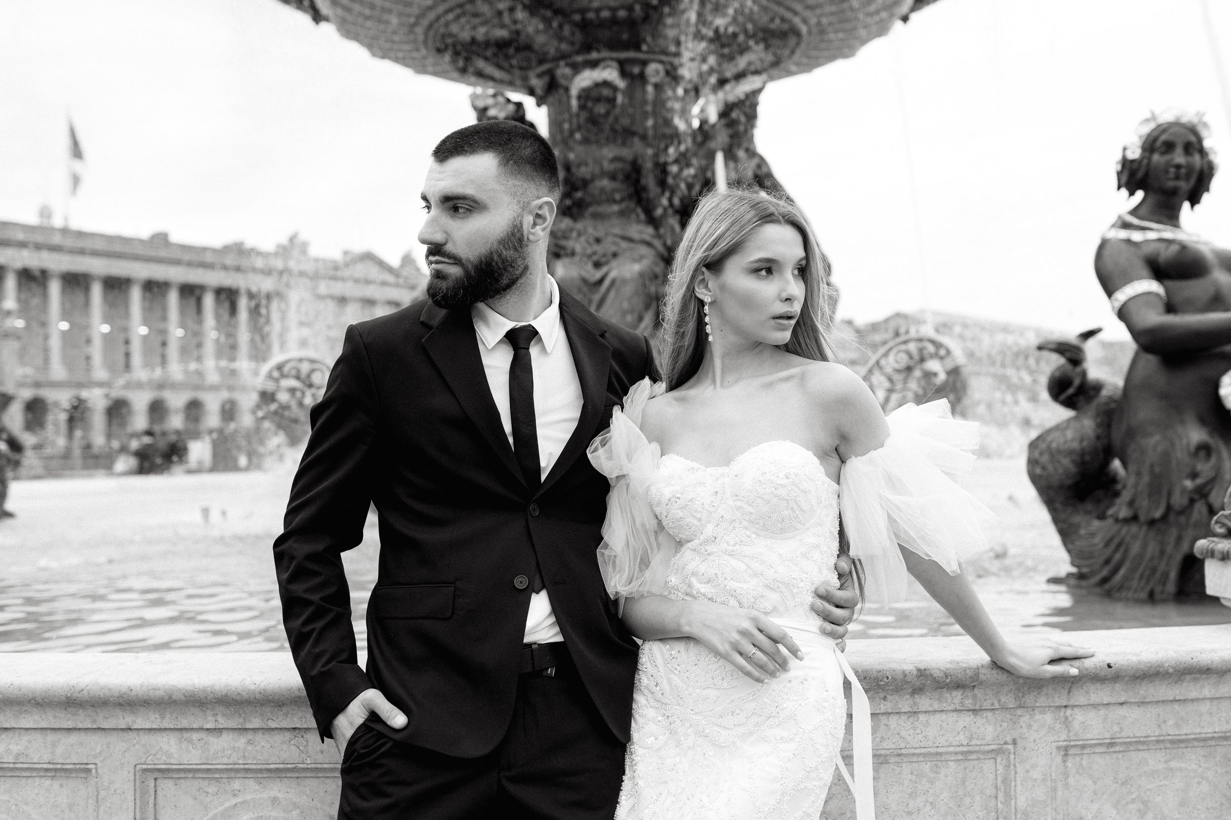 Un couple en tenue de mariage posant devant une fontaine en noir et blanc.