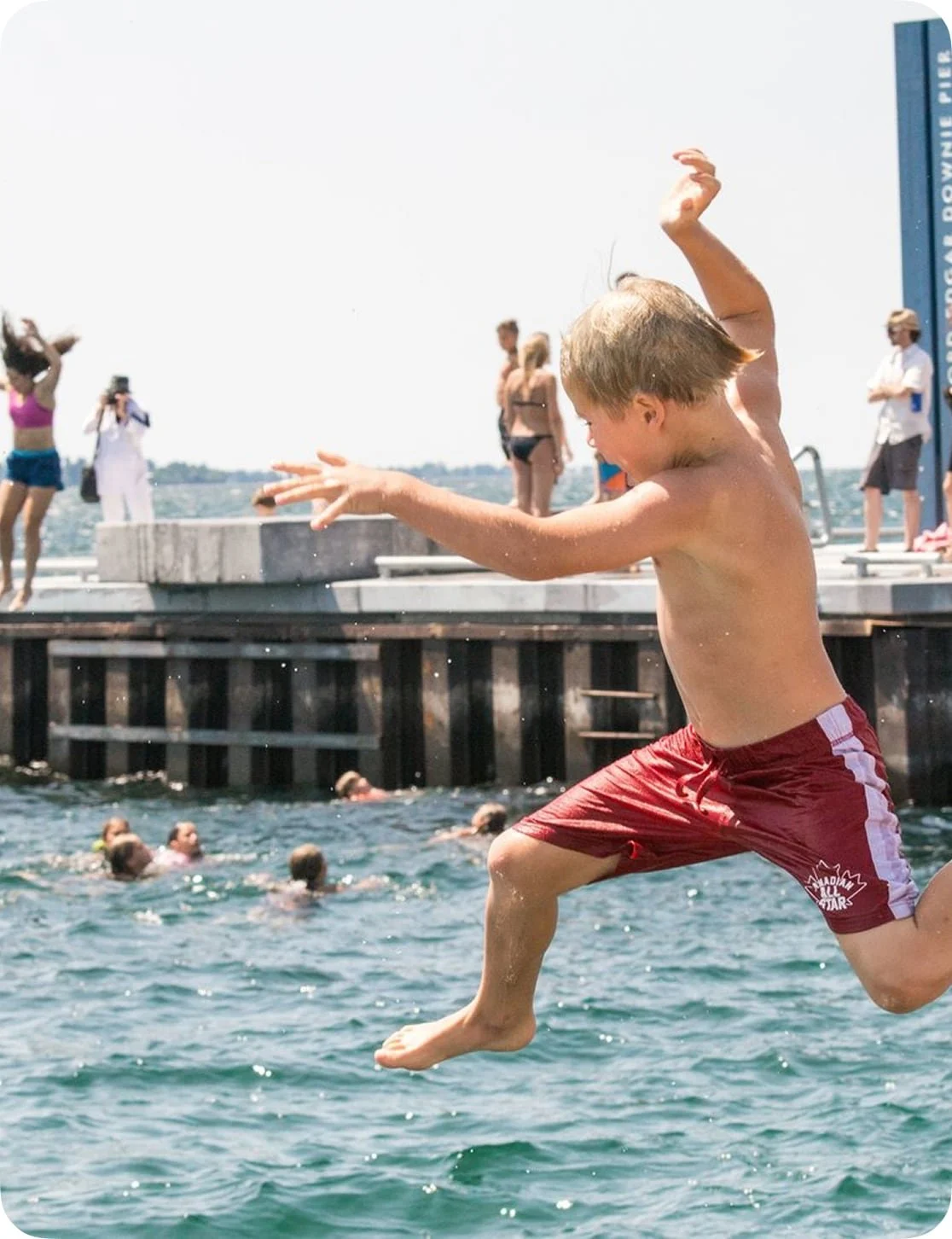 A young boy in red swim trunks jumping into the water from a dock on a sunny day, with people swimming and standing on the dock in the background.
