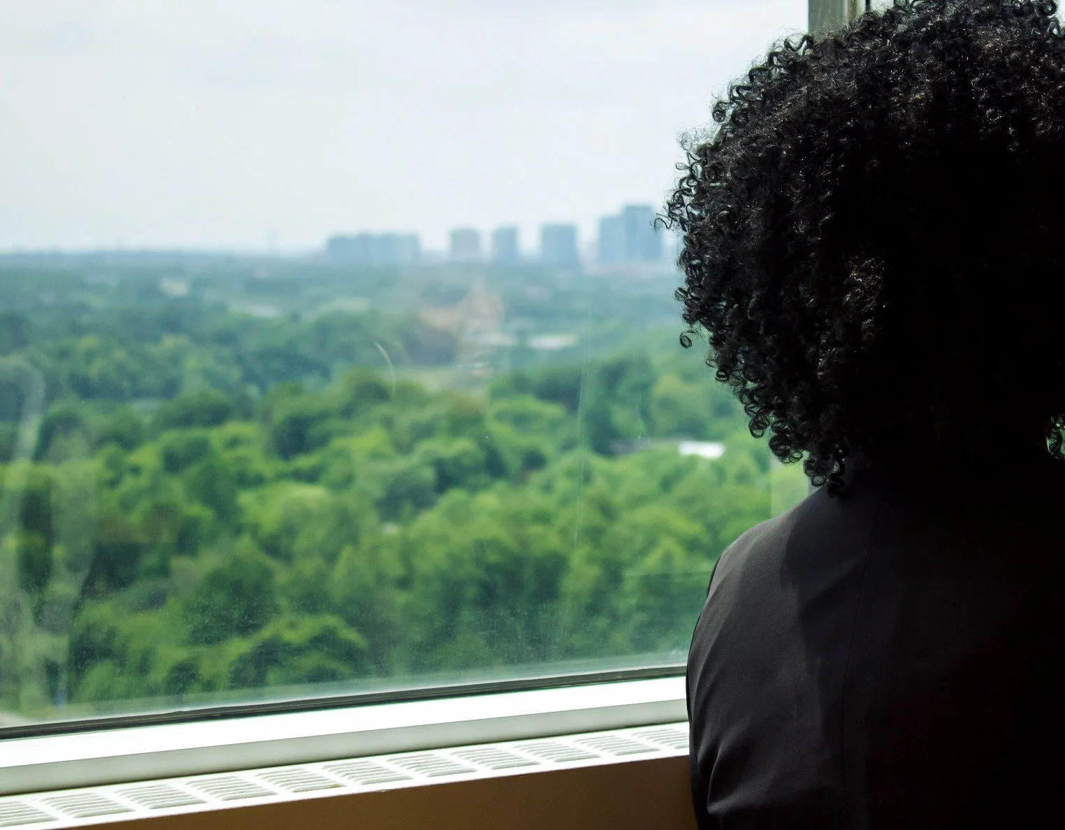 Person with curly hair looking out a window at a green landscape with city buildings in the distance.