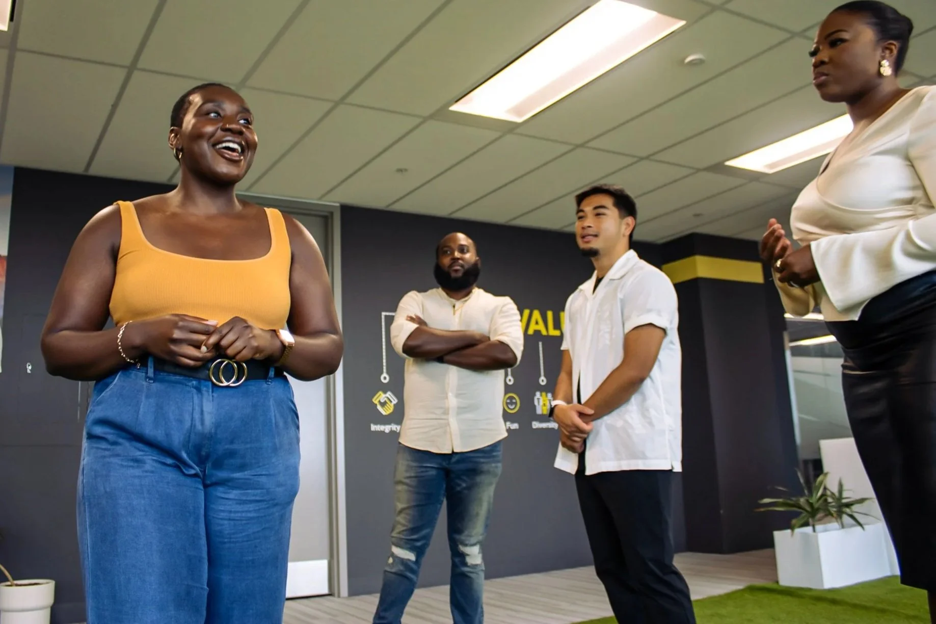 Four diverse professionals engaged in a discussion in a modern office. A woman in a yellow top and jeans is smiling, and three other colleagues are listening. The background features a wall with workplace values and a small plant.