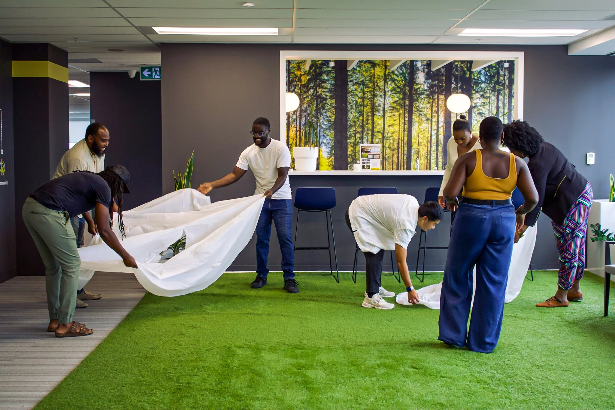 Group of people setting up over a green carpet in an indoor space, with a nature-themed wall art in the background.