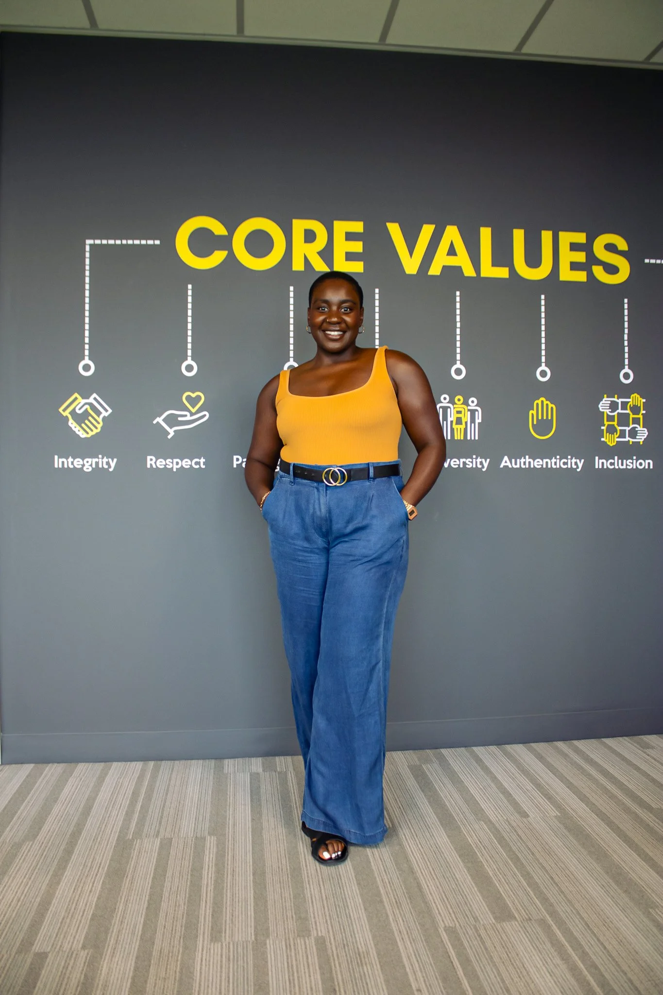A woman standing in front of a wall displaying core values, smiling and wearing a yellow sleeveless top and blue wide-leg pants.