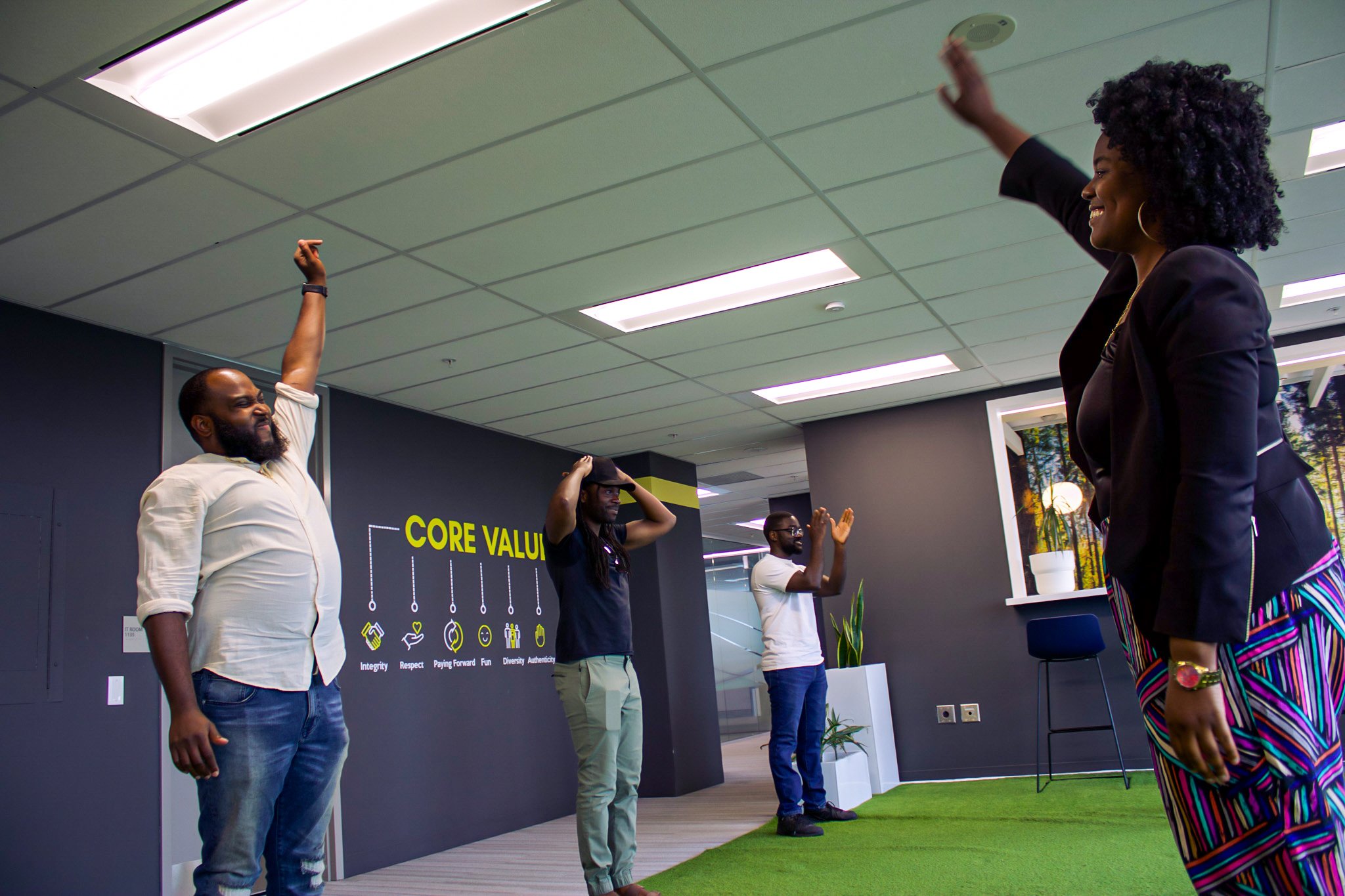 Four diverse colleagues participate in a team-building or training activity in an office, with one woman and three men standing apart, raising their arms or clapping, against a wall displaying company core values.